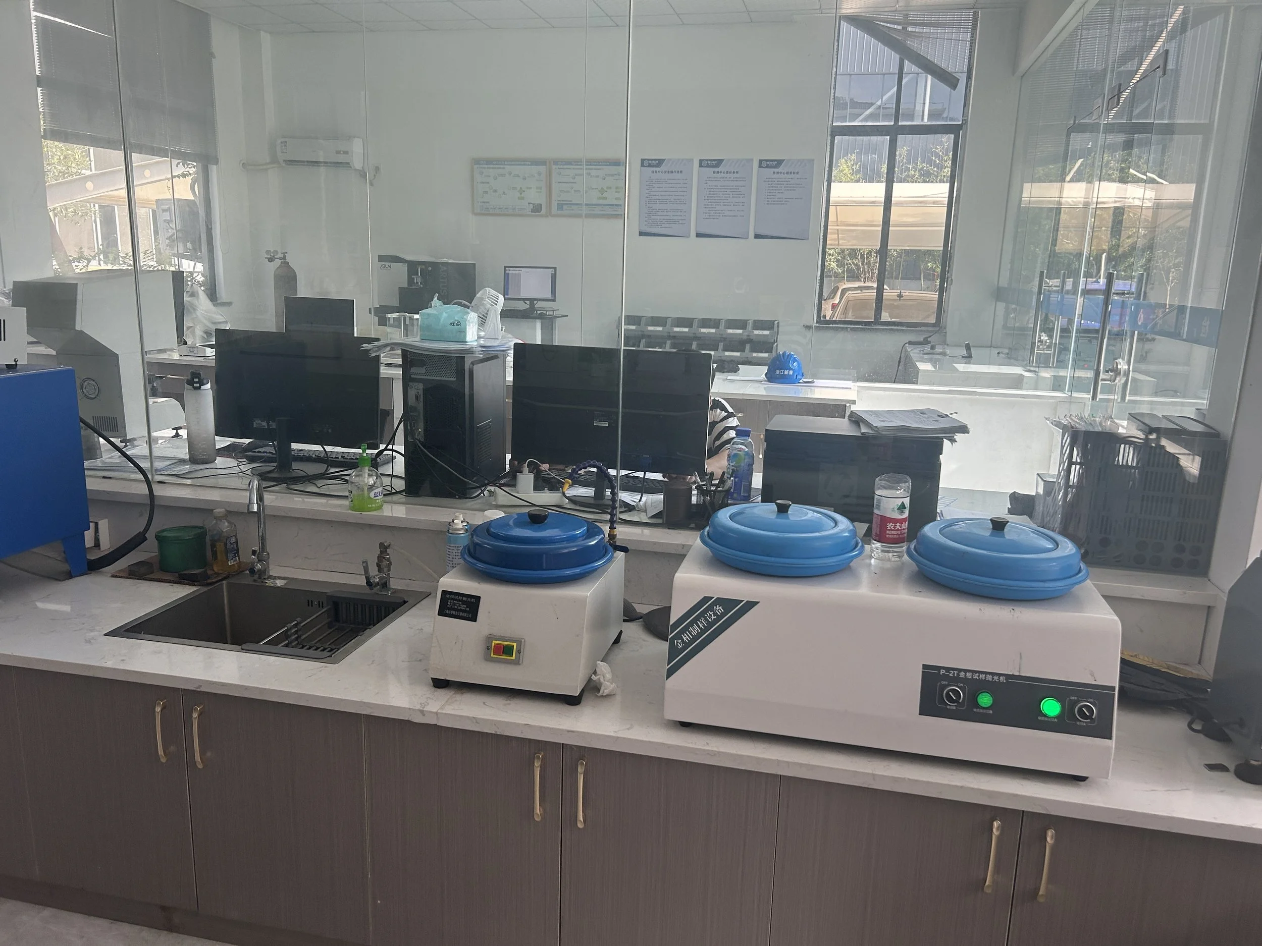 Laboratory kitchen with two blue-lidded sterilization machines on the counter, a sink, and office equipment visible behind a glass partition.