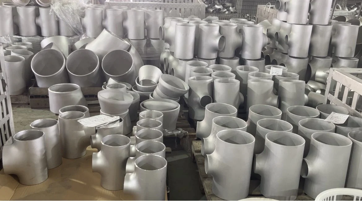 A large warehouse filled with numerous Stainless steel pipe fittings, including elbows and tees, arranged on pallets and shelves.