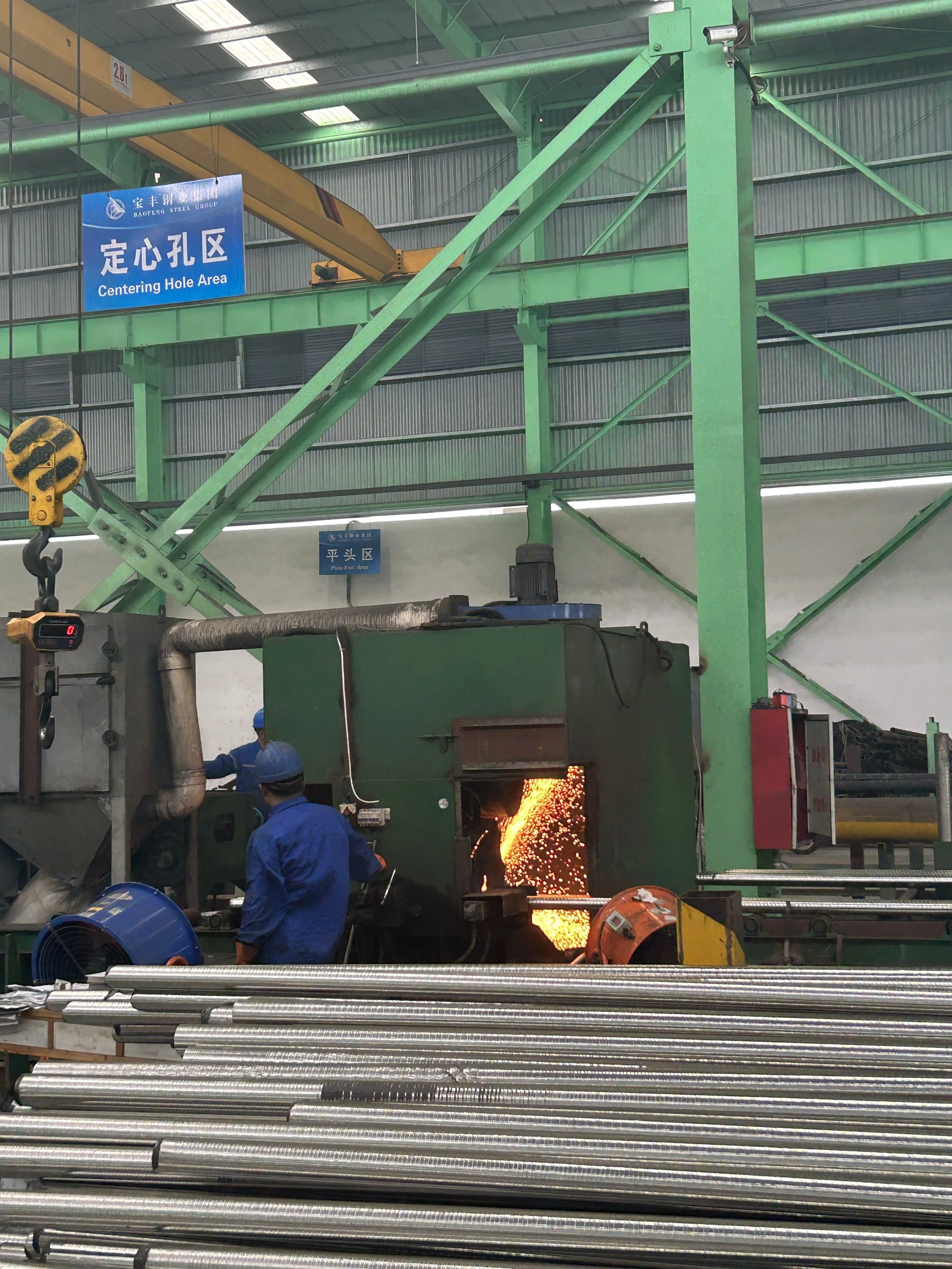 Workers use a machine with sparks in an industrial steel manufacturing or processing plant with green structural beams, signboards in Chinese and English, and metal pipes in the foreground.