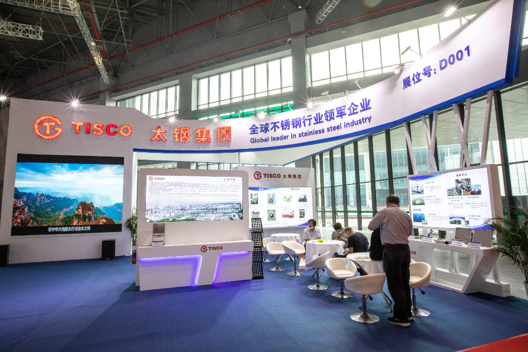 Trade show booth for TISCO Steel Group with large screens showcasing steel industry information, and seating area with visitors and representatives, inside a spacious exhibition hall with high ceiling and large windows.