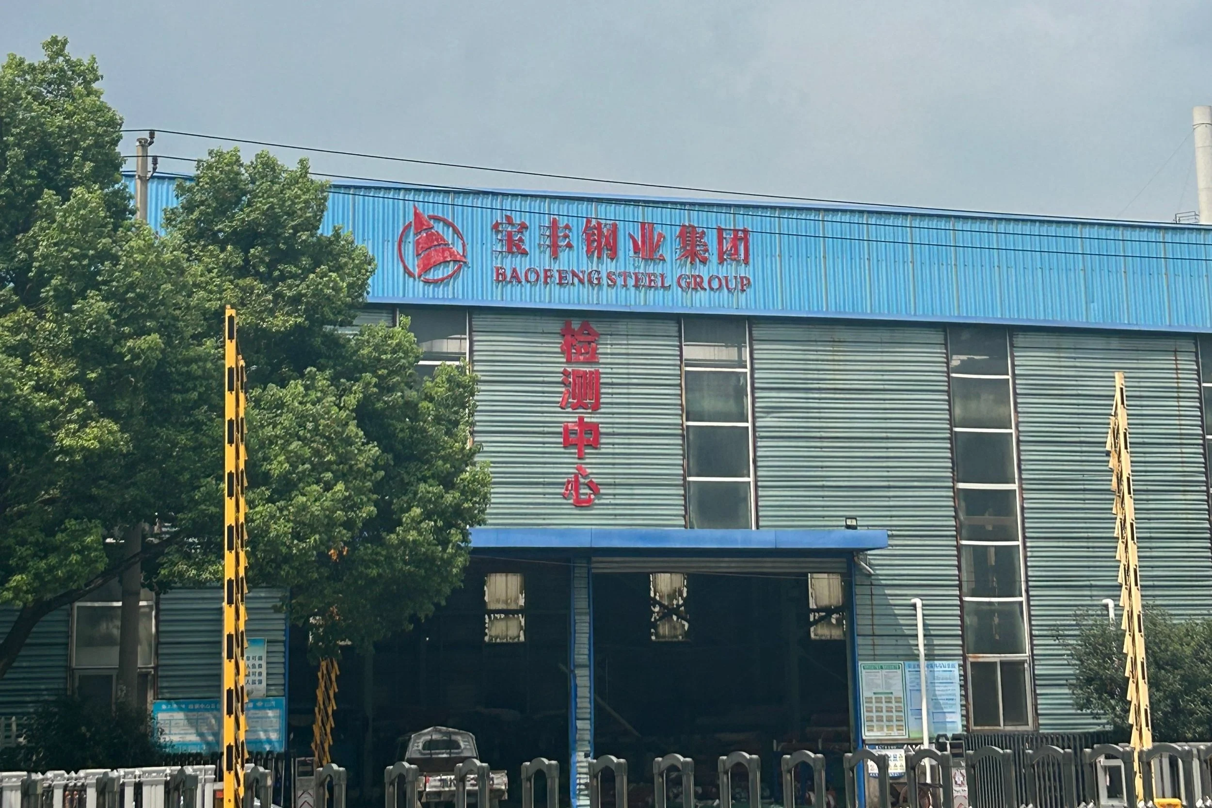 Industrial building with blue siding, red signage displaying 'Baofeng Steel Group' and Chinese characters, green trees in front, and a parking area.