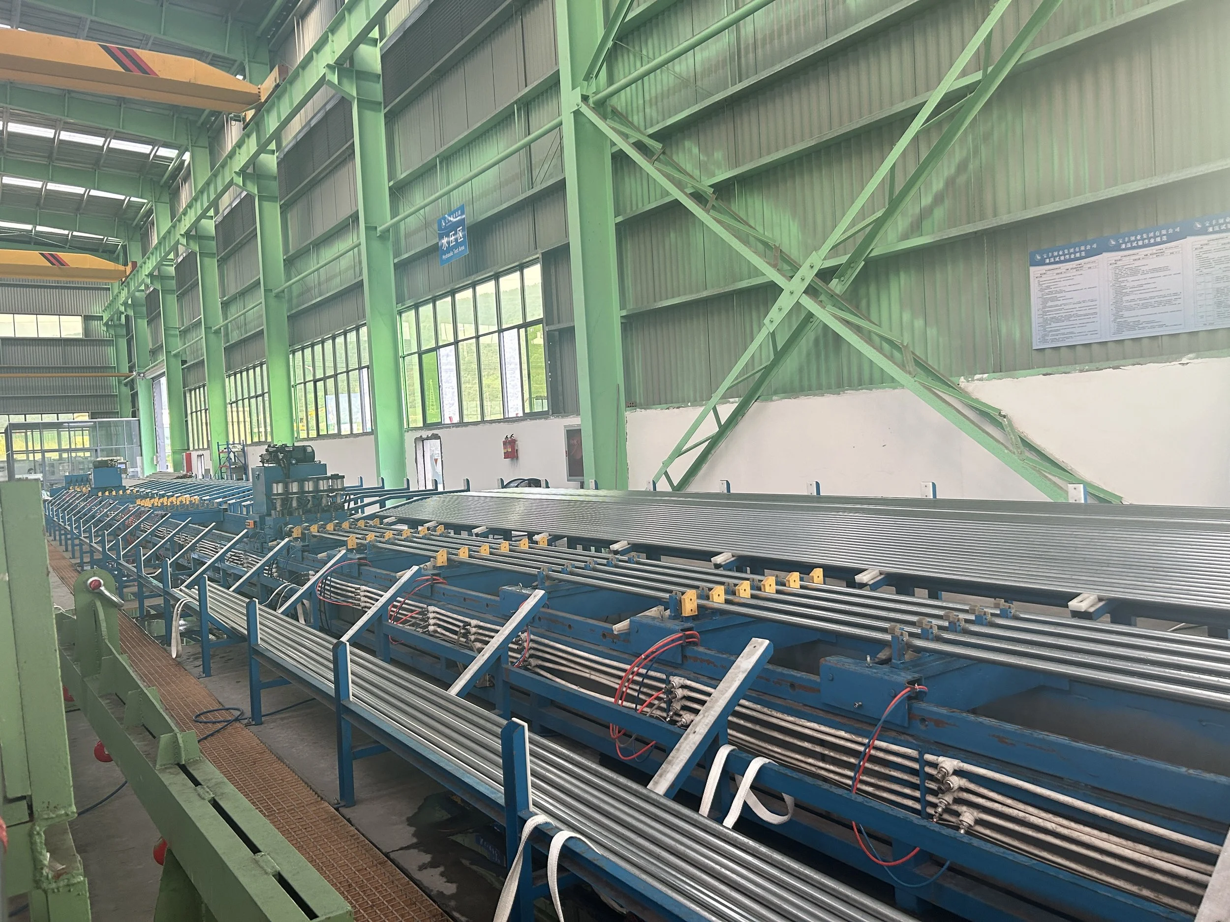 Interior of an industrial manufacturing facility with green metal support beams, large windows, and a blue automated conveyor system for transporting materials.