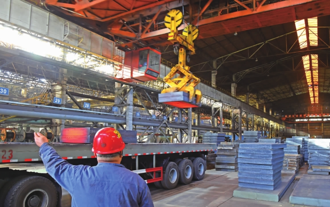 A worker wearing a red safety helmet and blue uniform is gesturing inside a large industrial warehouse. The warehouse has a high ceiling with infrared lighting and is filled with steel beams, machinery, and stacks of steel plates. A yellow overhead crane is lifting a large steel plate.