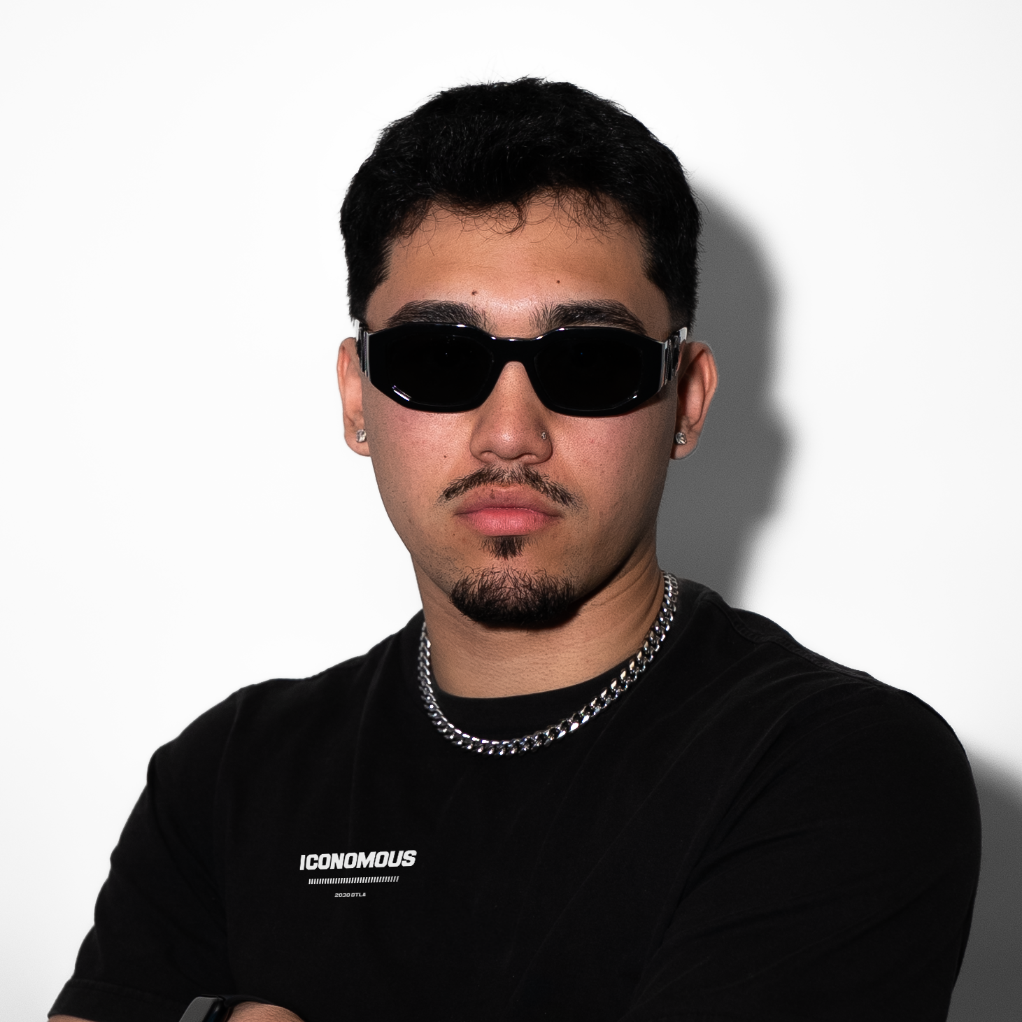 A young man with black hair, wearing black sunglasses, a black T-shirt with white text, and a silver chain necklace, standing against a white background.