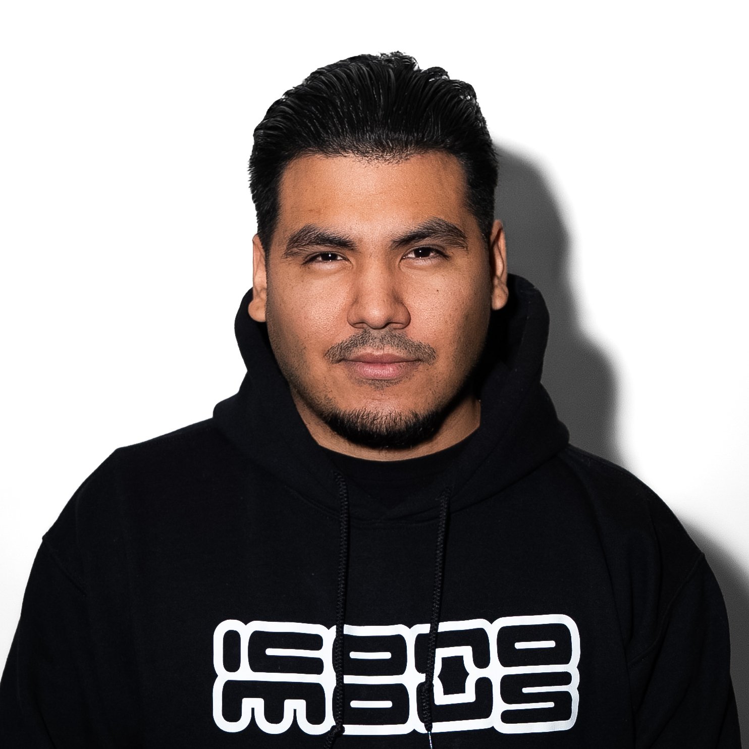 A man with dark hair and facial hair wearing a black hoodie with white text, standing against a white background with a shadow cast on the wall.