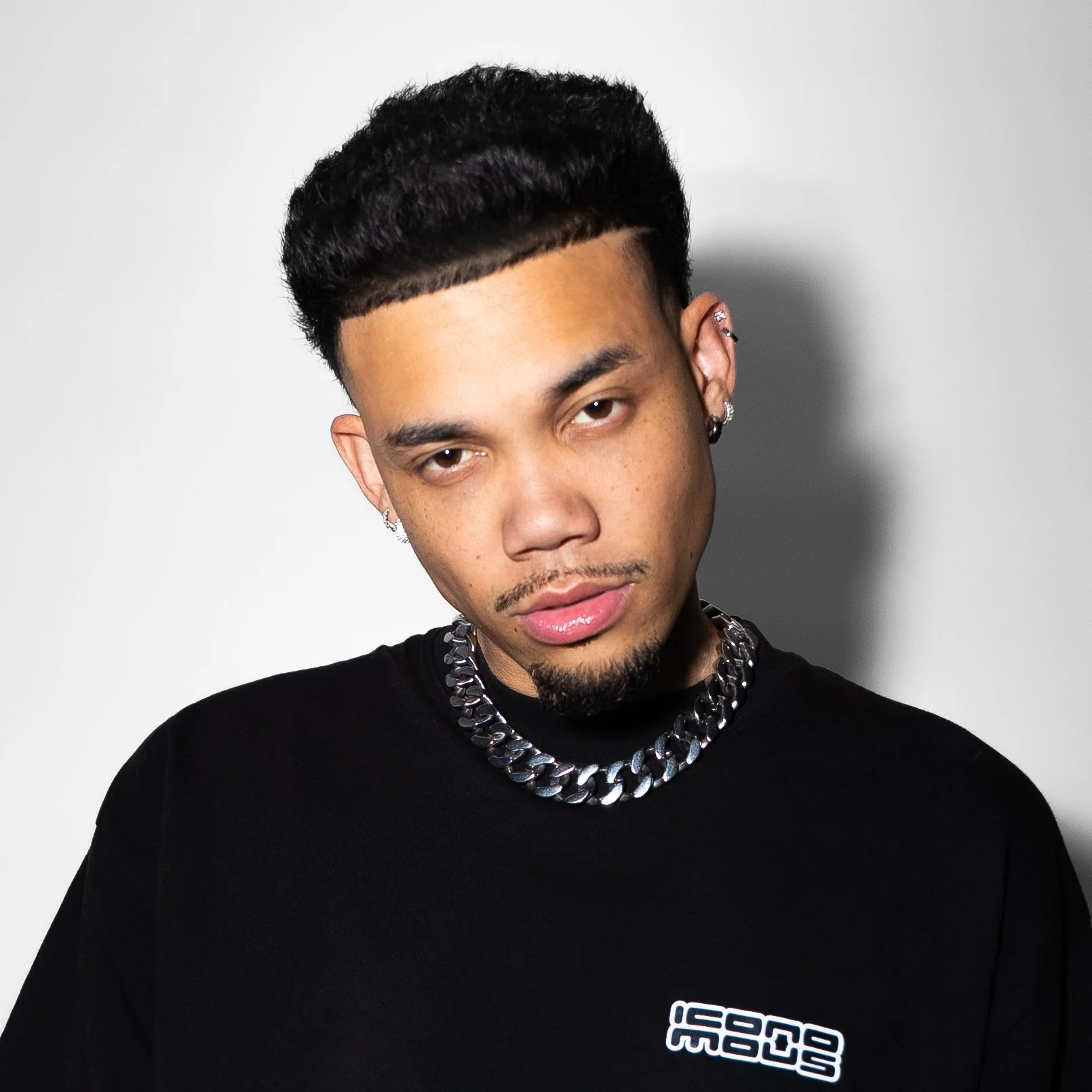 Roshon Fegan with short black hair, multiple earrings, wearing a black t-shirt with white text and a chunky silver chain necklace, looking at the camera against a plain white background.