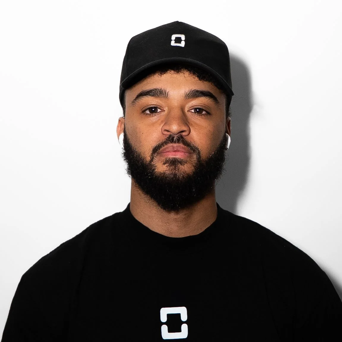 A young man with a beard, wearing a black baseball cap and black shirt, looking directly at the camera, white background.