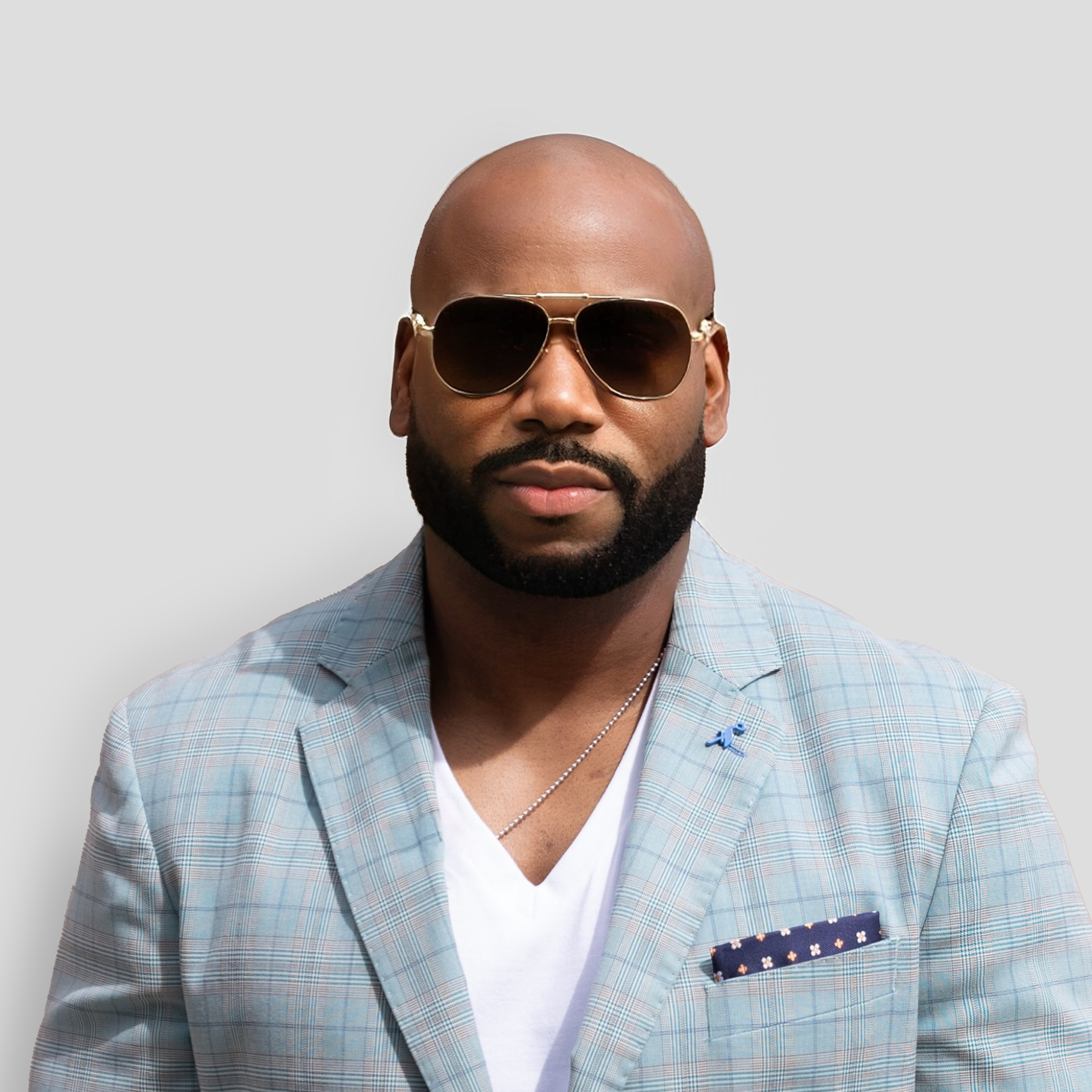 A man with a shaved head, beard, wearing sunglasses, a light-colored blazer, and a white V-neck shirt, standing against a plain background.