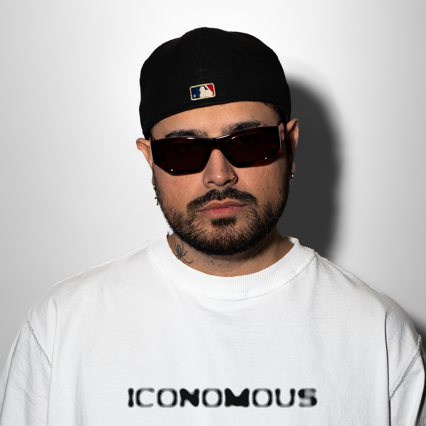A man wearing sunglasses, a black cap with a sports league logo, a nose ring, earrings, a tattoo on his neck, and a white sweatshirt with the word 'ICONOMOUS' printed on it.
