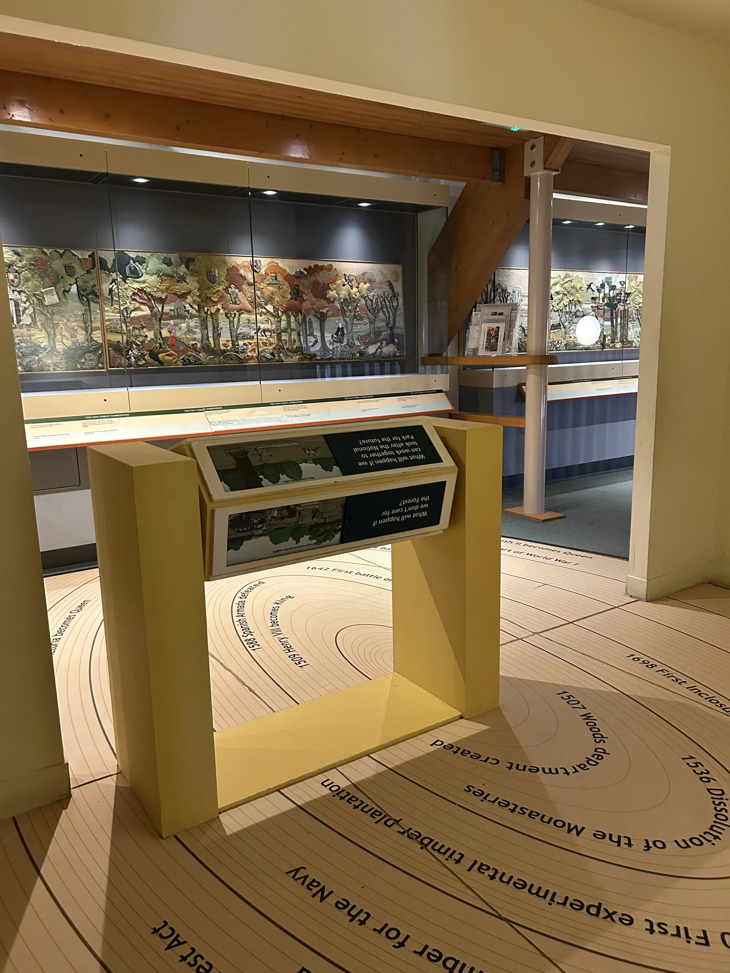 Exhibit of embroidered tapestry in a museum display case, with orange and white informational stand in the foreground.