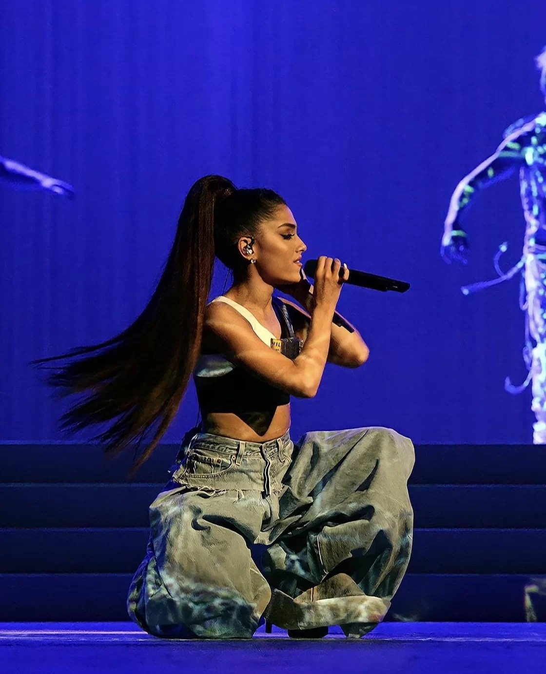 Performer wearing high ponytail and baggy jeans, holding a microphone on stage with blue lighting.