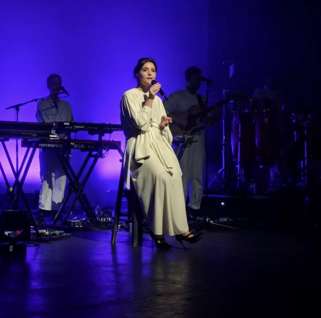 Singer performing on stage with band, wearing white dress, purple lighting.