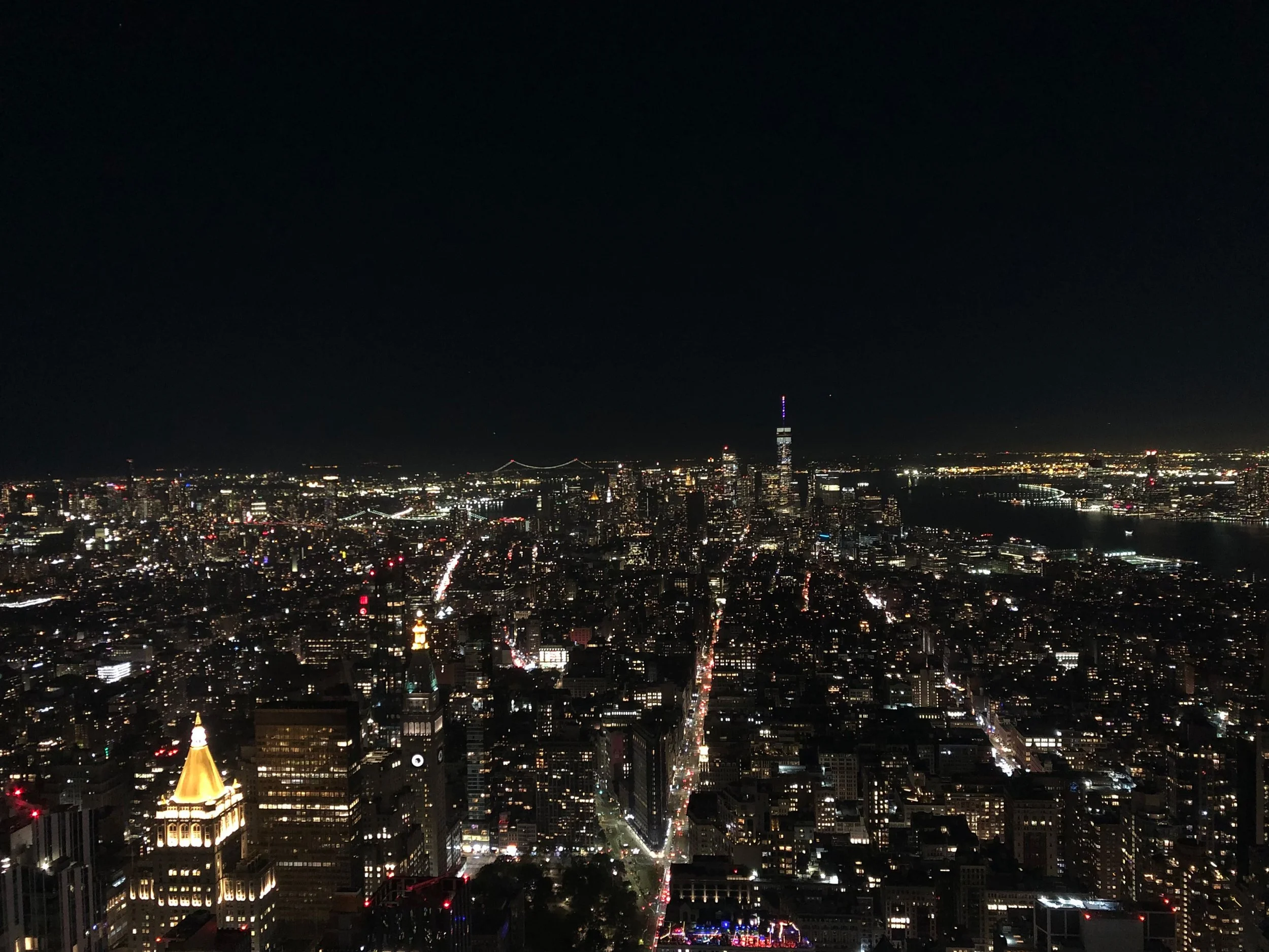 Aerial night view of New York City skyline with illuminated buildings and streets, featuring the Hudson River and prominent skyscrapers.