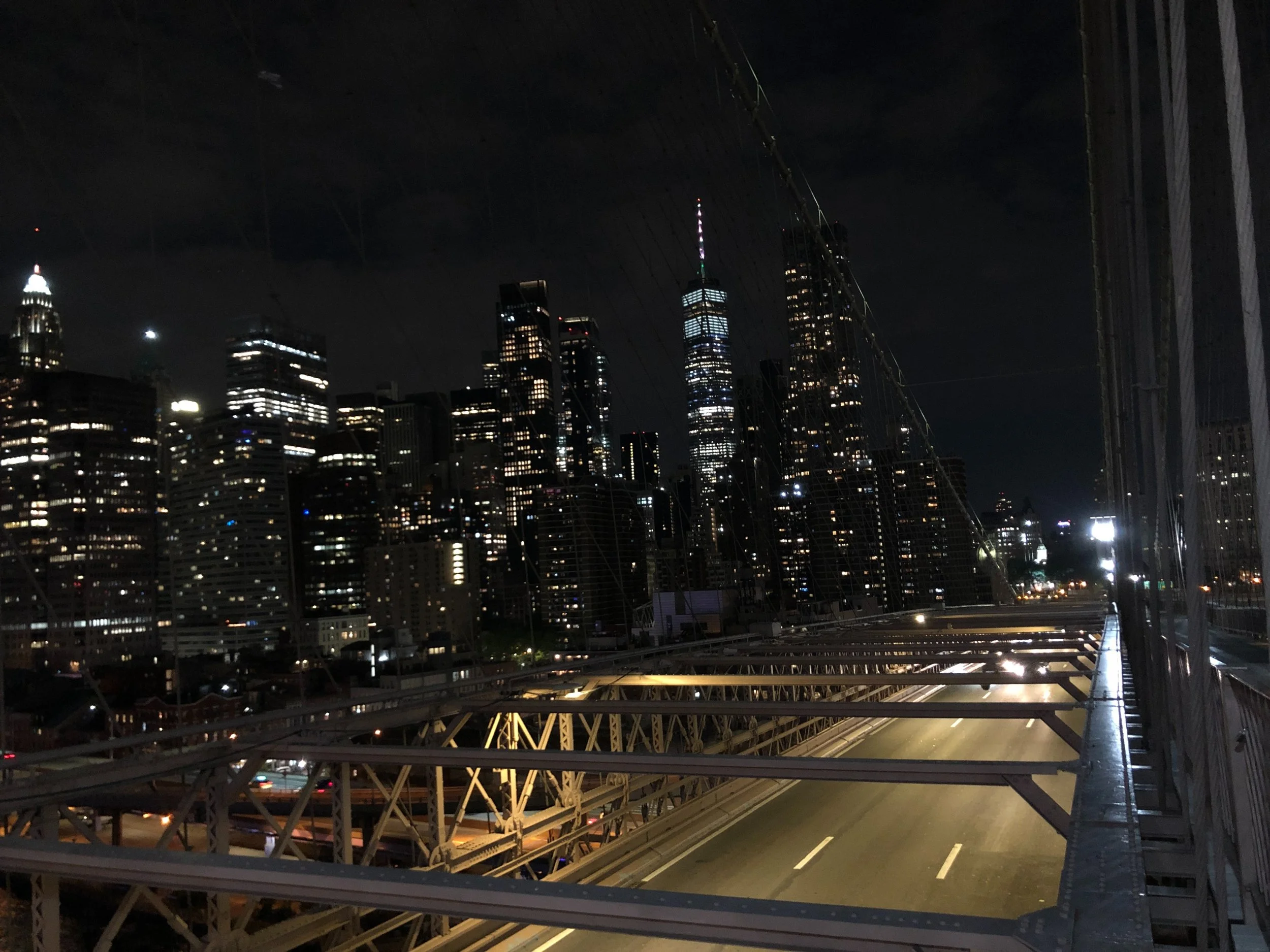New York City skyline at night with illuminated skyscrapers and a view of a highway bridge, possibly the Brooklyn Bridge. The One World Trade Center is visible in the background.
