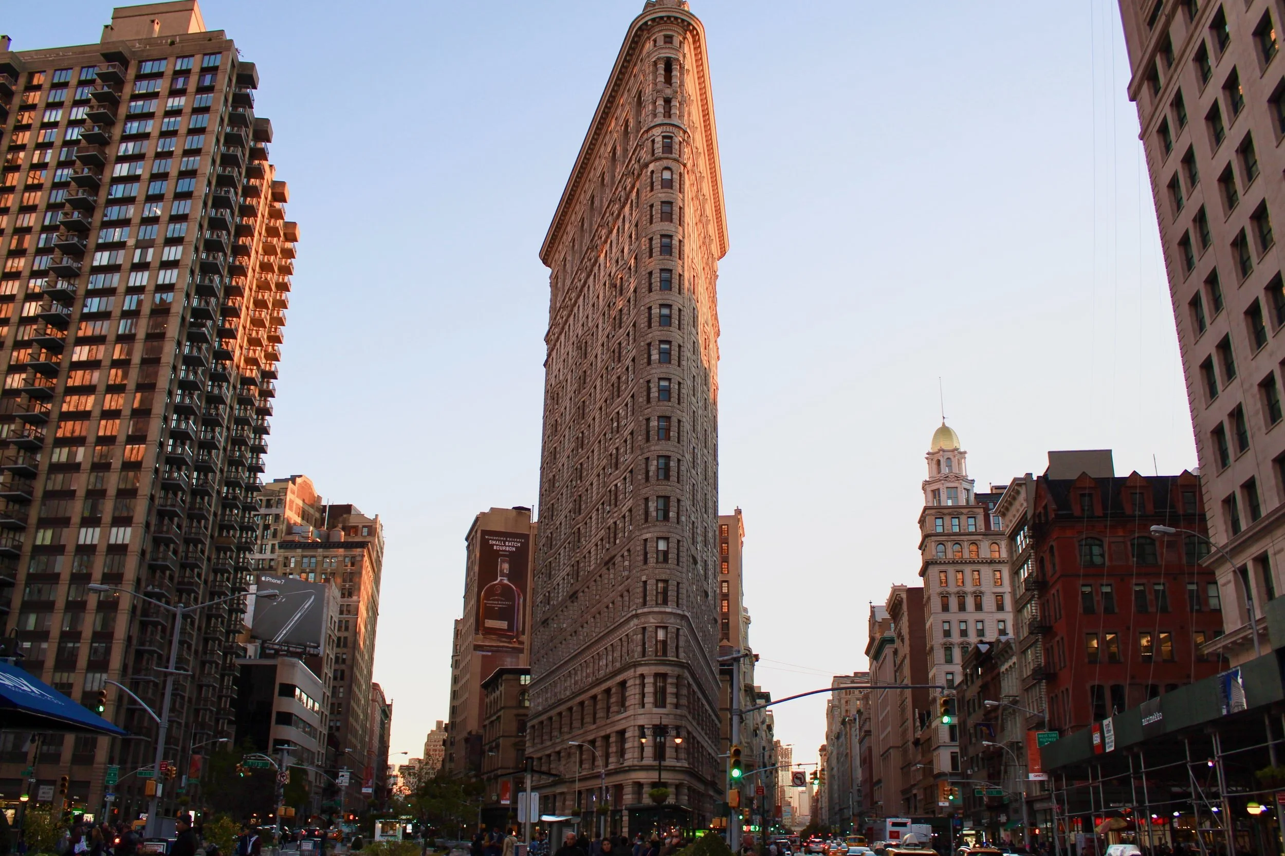 Flatiron Building in New York City at sunset, surrounded by other tall buildings and street traffic.