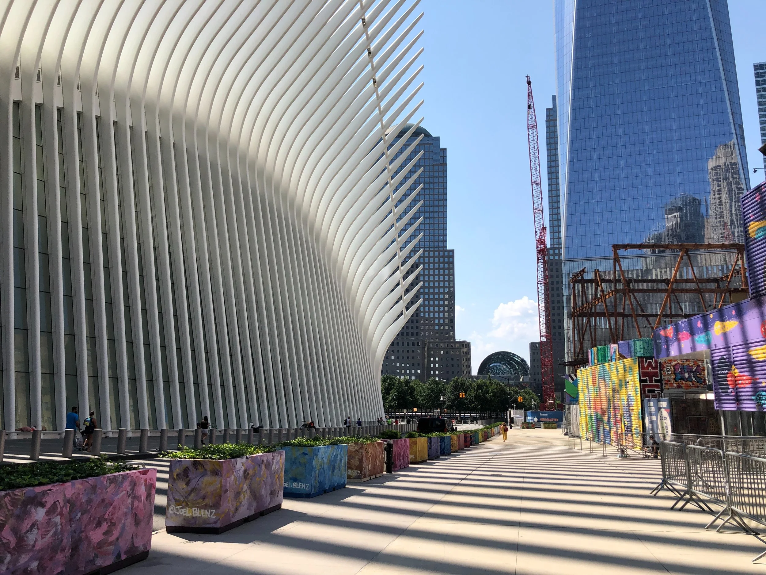 Modern architectural structure with curved white ribbed design, adjacent to a tall glass skyscraper, and colorful painted panels in an urban setting.