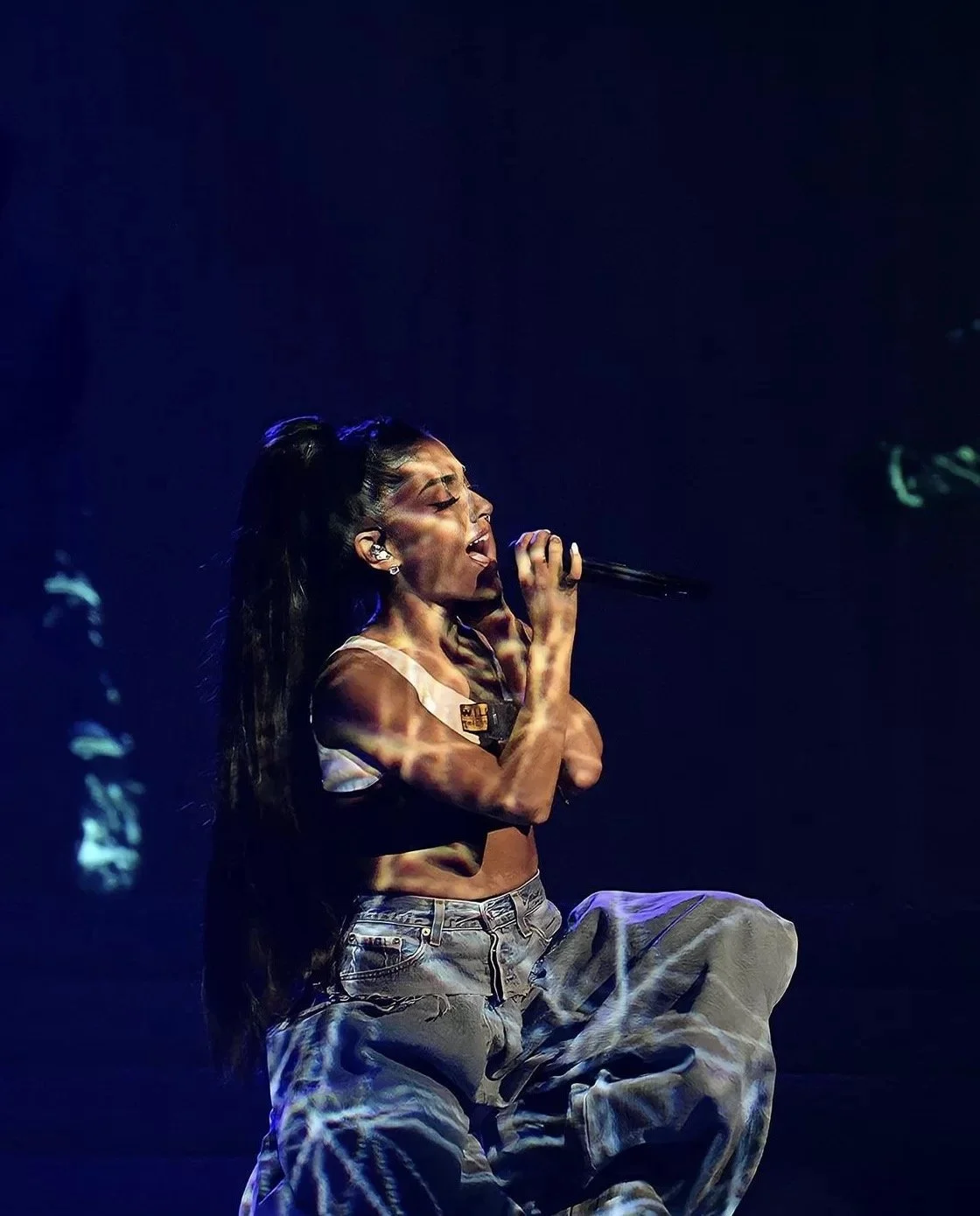 Performer singing energetically on stage in casual attire, including a white top and baggy jeans, holding a microphone under dramatic lighting.