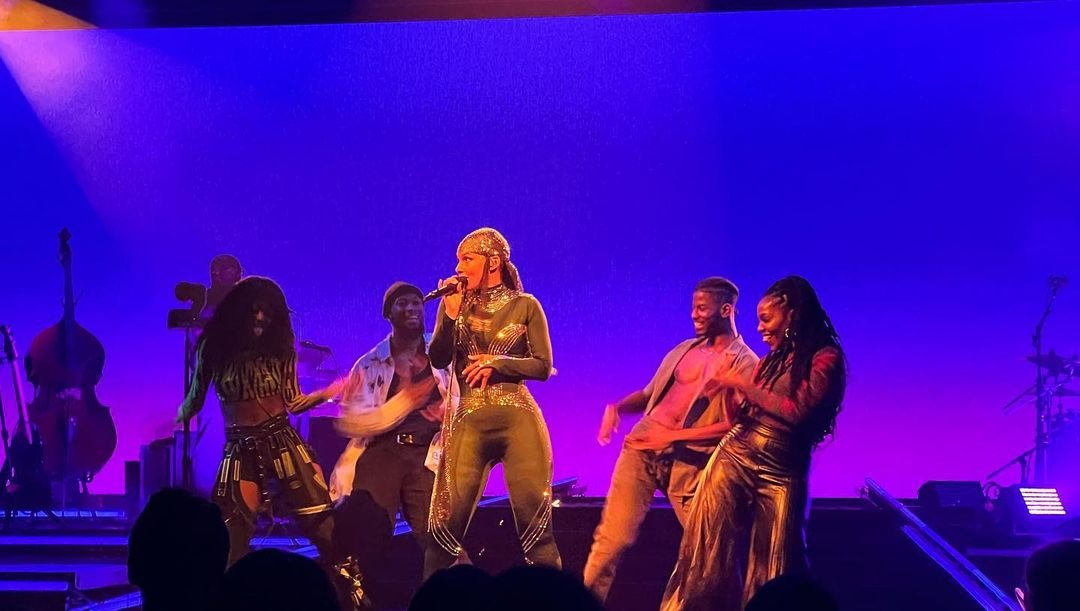 Performer singing on stage with dancers and musicians, purple background, dynamic lighting.