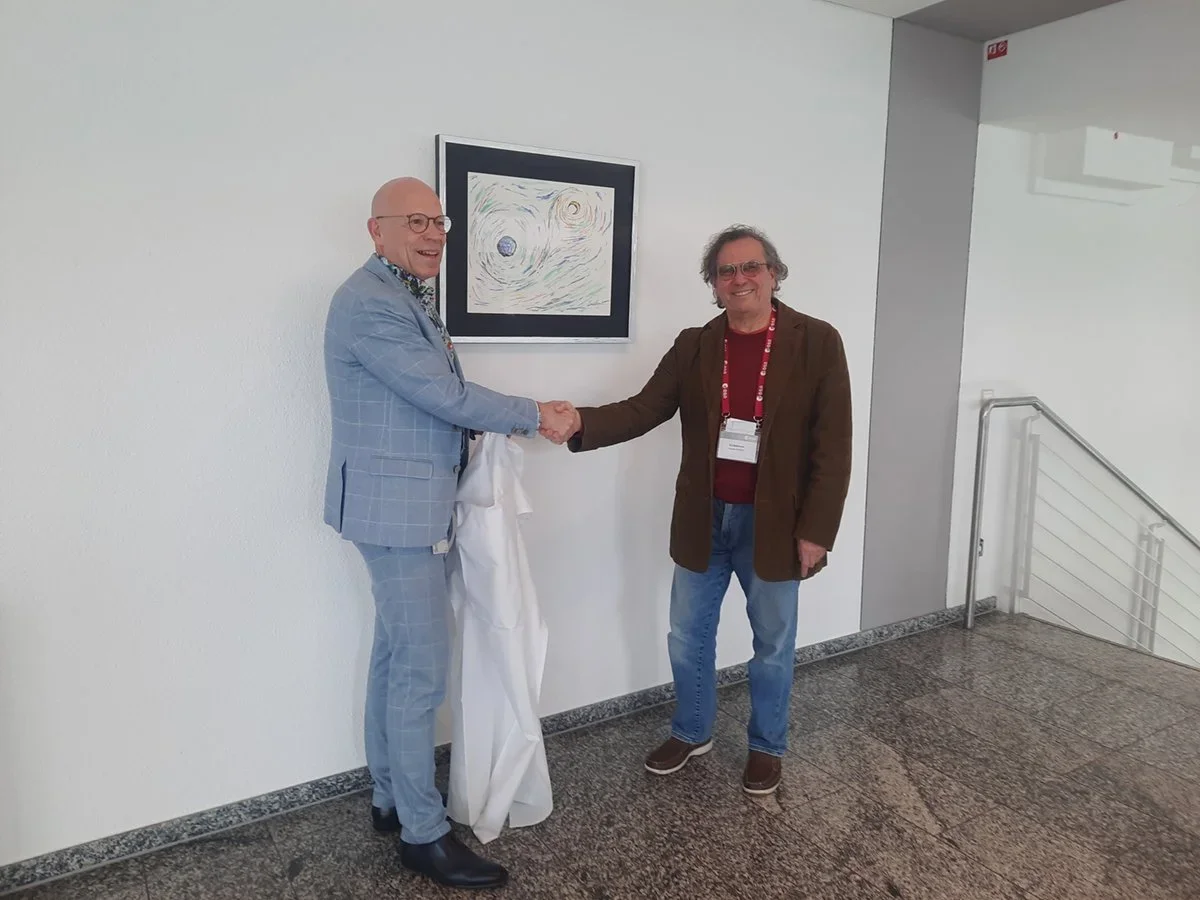 Two men shaking hands in a hallway, one in a light blue suit and glasses, the other in a brown jacket and glasses. They are near a framed artwork on the wall.
