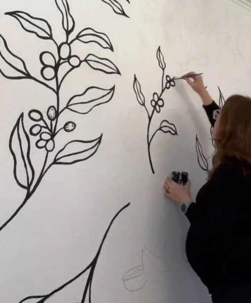 Person drawing large black outlines of coffee plant with berries and leaves on a white wall.