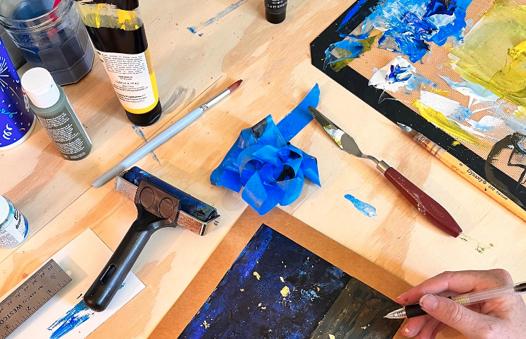 Artist's workspace with acrylic paints, brushes, and tools on a wooden table, including a palette with blue and yellow paint, a blue paper flower, and a partially painted canvas.