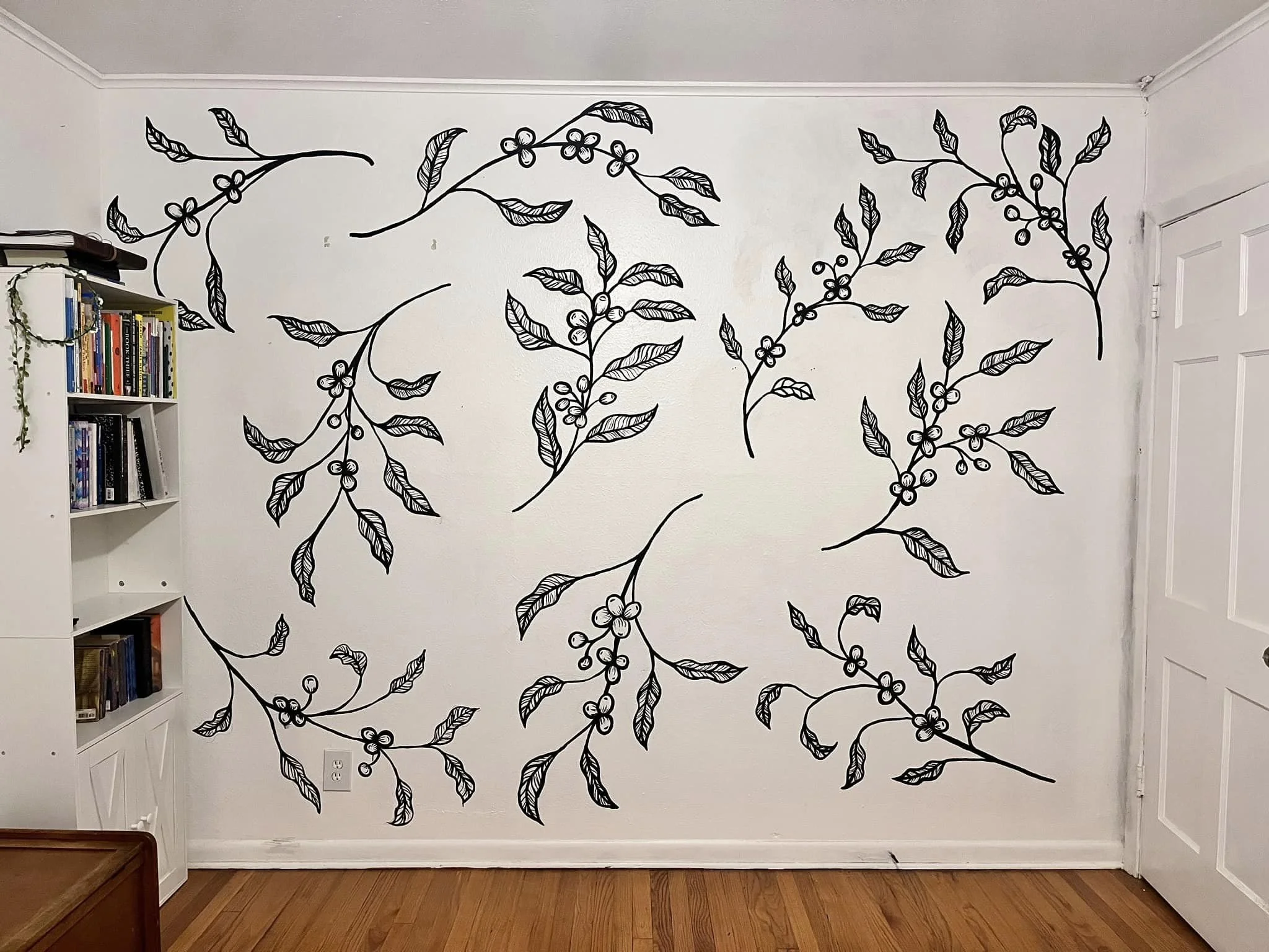Black outline drawing of coffee plant branches with leaves and berries on a white wall in a room.