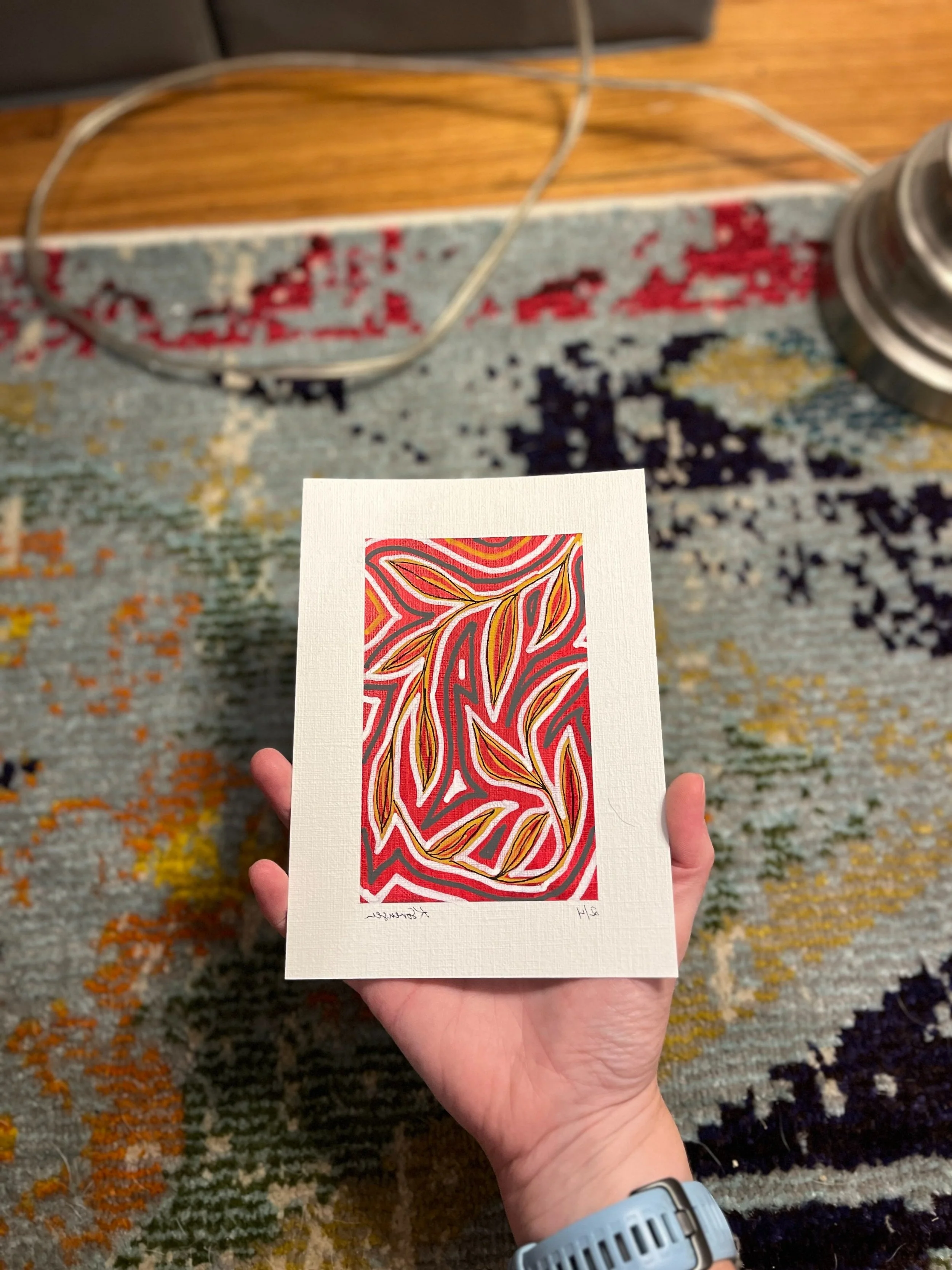 A person is holding a small piece of artwork with an intricate, colorful, abstract pattern of swirling lines and leaf shapes in red, yellow, white, and black on a white background, with some handwritten text below.