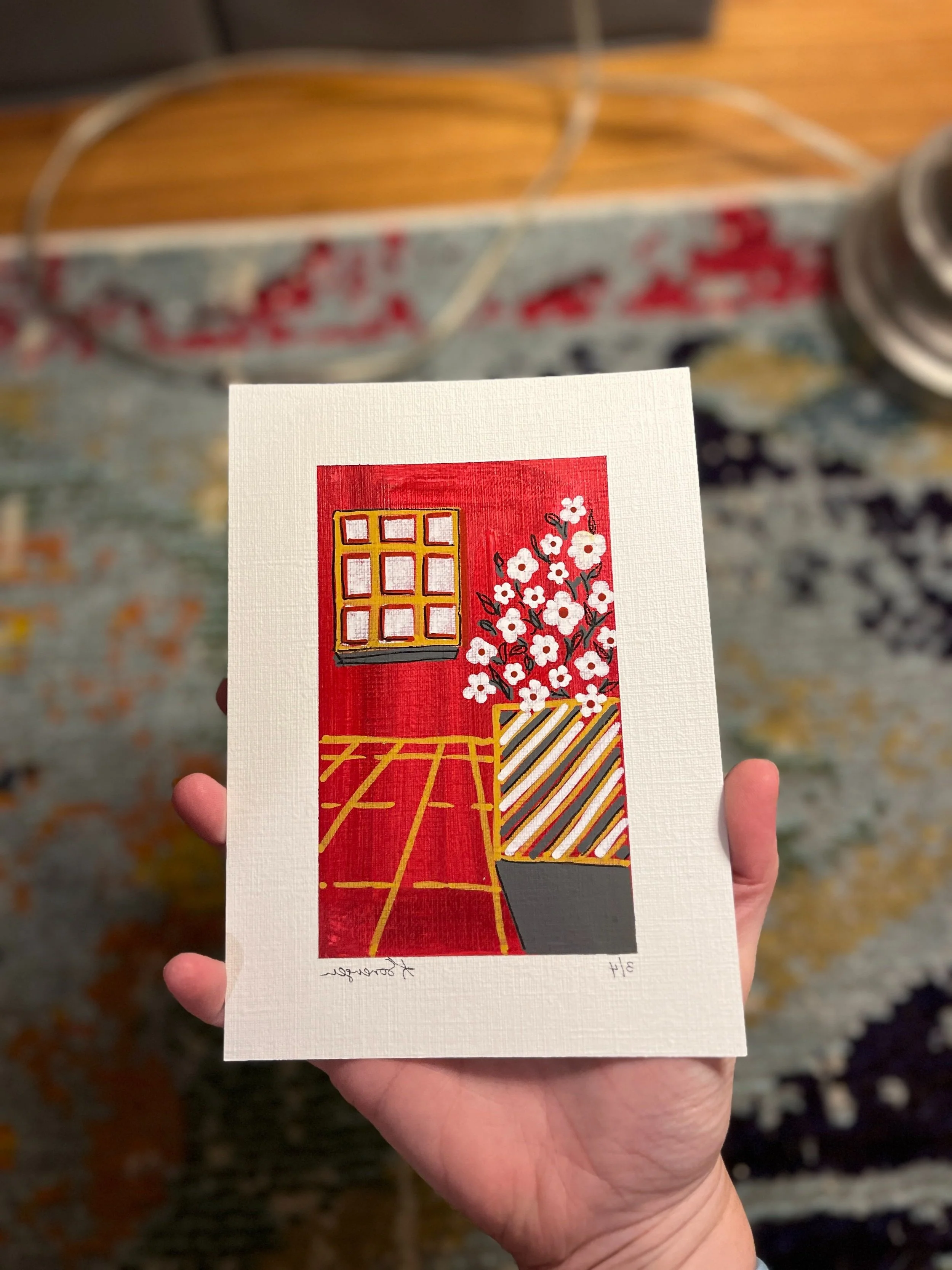 A hand holding a card with a painting of a red room featuring a window, a potted plant with white flowers, and a patterned vase. The background shows a blurred colorful rug and part of a vacuum cleaner.