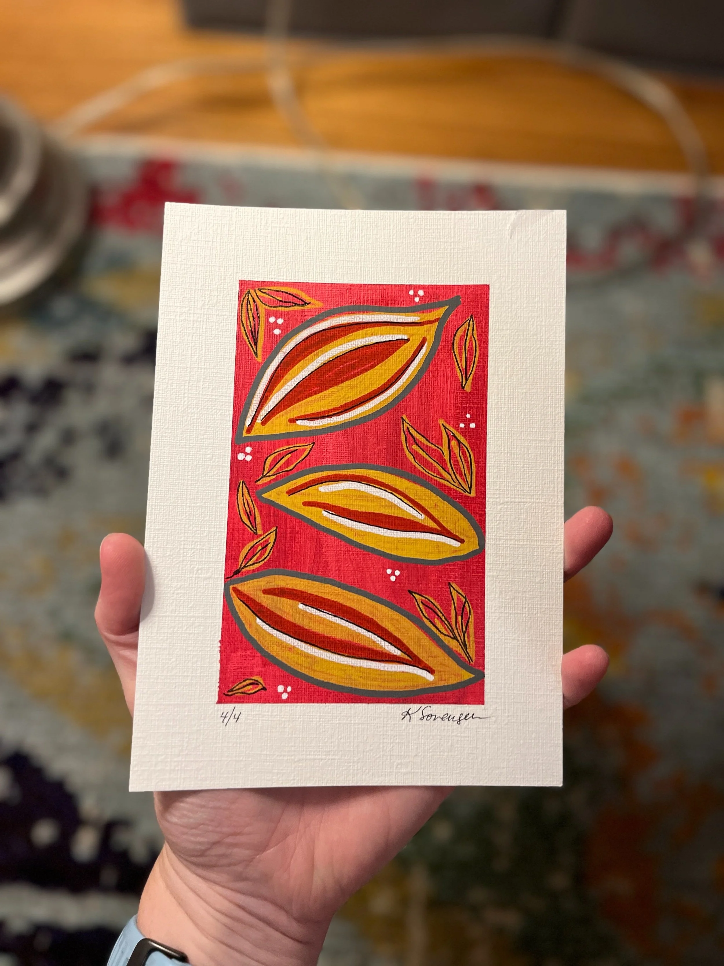 A small, colorful print of abstract leaves on a red background, held in a person's hand.