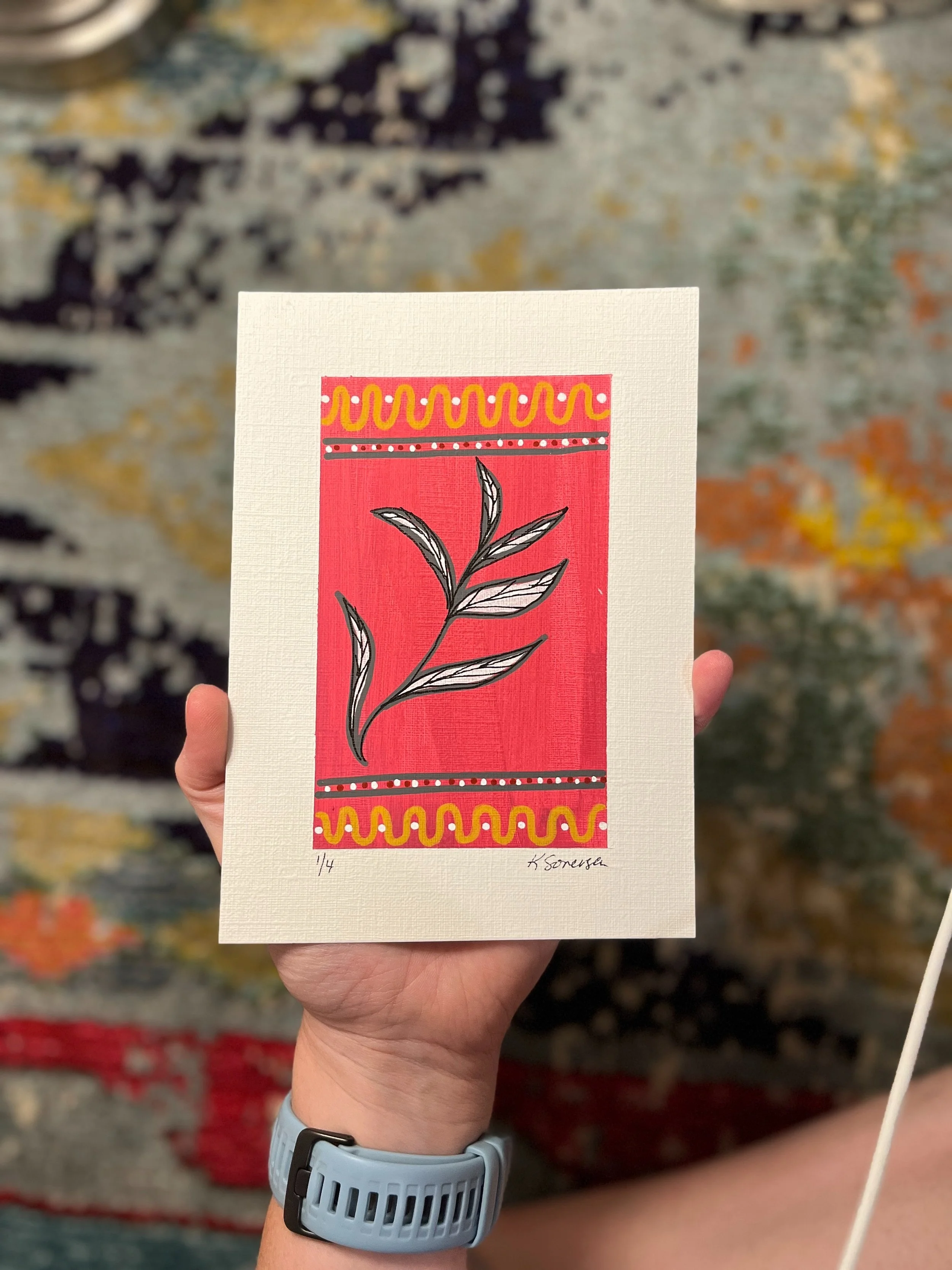 A person holding a colorful art print featuring a black and white illustrated branch with leaves on a red background. The print has decorative yellow and pink borders at the top and bottom, and is signed by the artist.