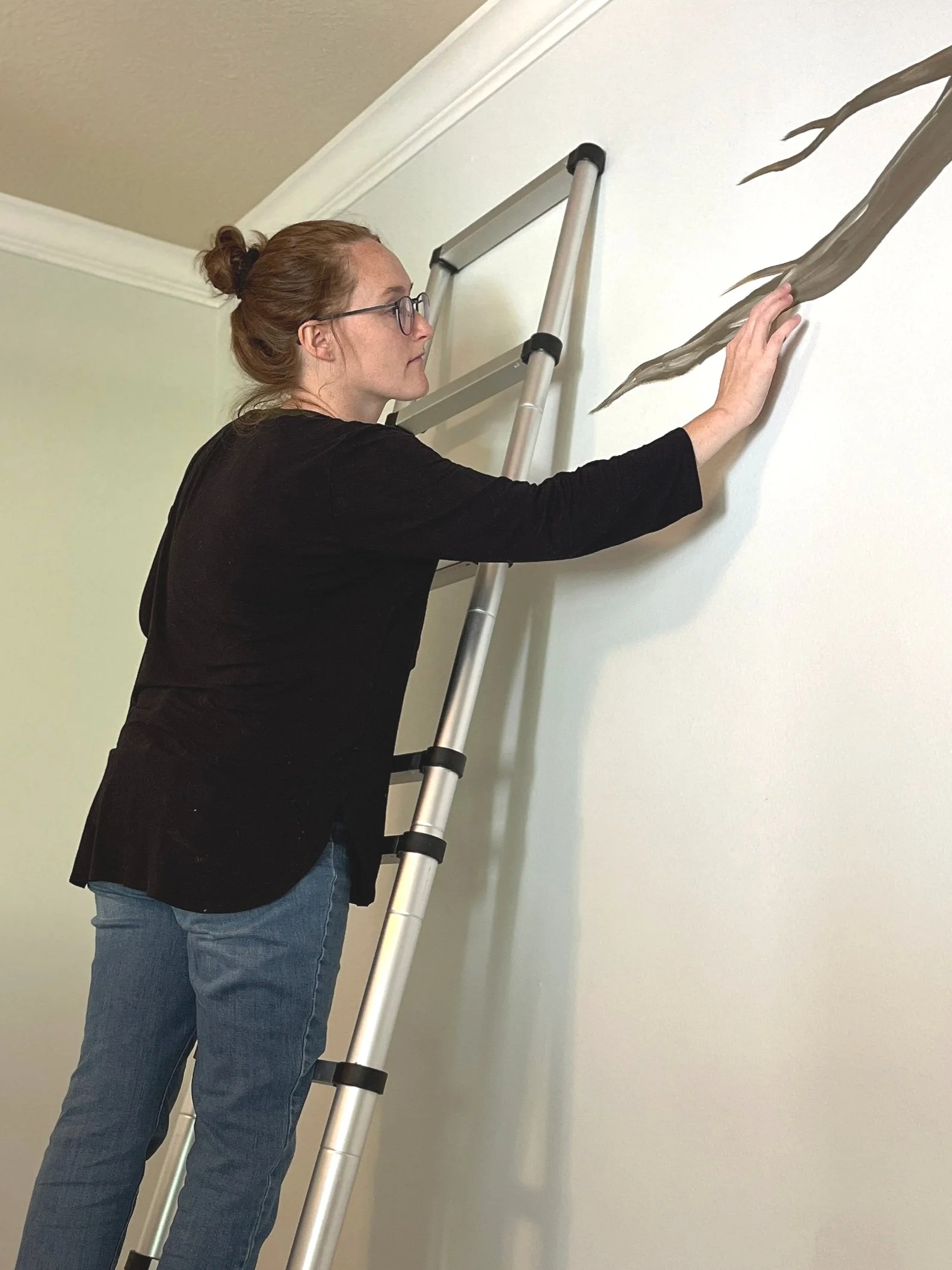 Woman standing on a ladder painting a vine or branch on the wall.