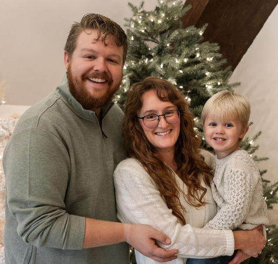 A family of three smiling and posing together in front of a decorated Christmas tree, indoors.