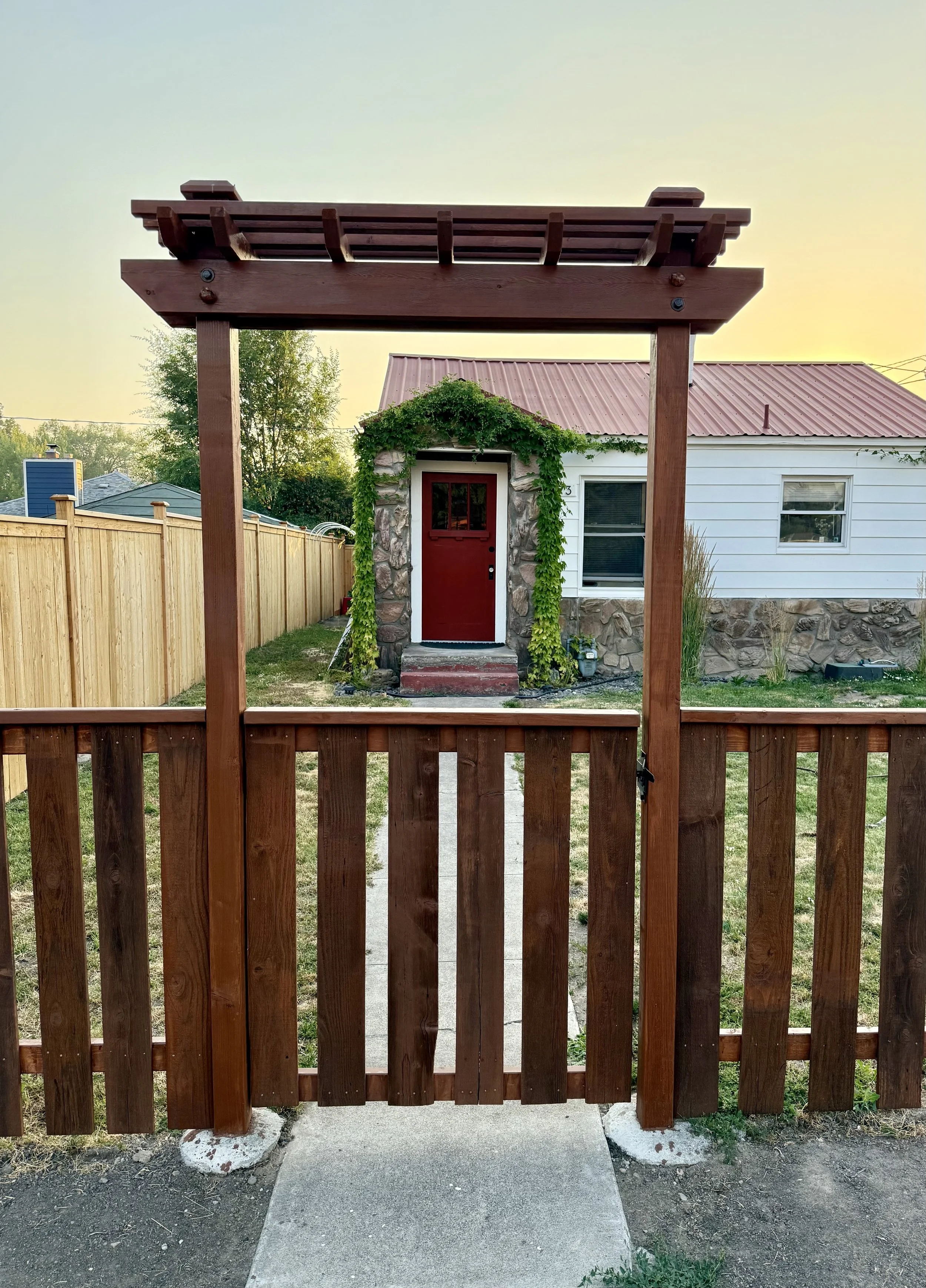 View of a backyard gate framing a small house with a red door, ivy-covered wall, and a red metal roof, with a wooden fence on the left side and a white house in the background.