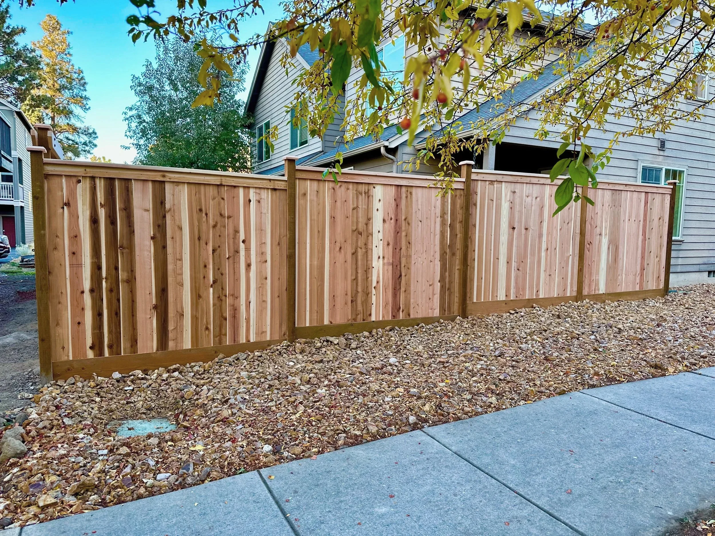 A newly installed wooden privacy fence along a residential property, with a gravel ground cover and a concrete sidewalk in the foreground. There are trees and a multi-story house in the background.
