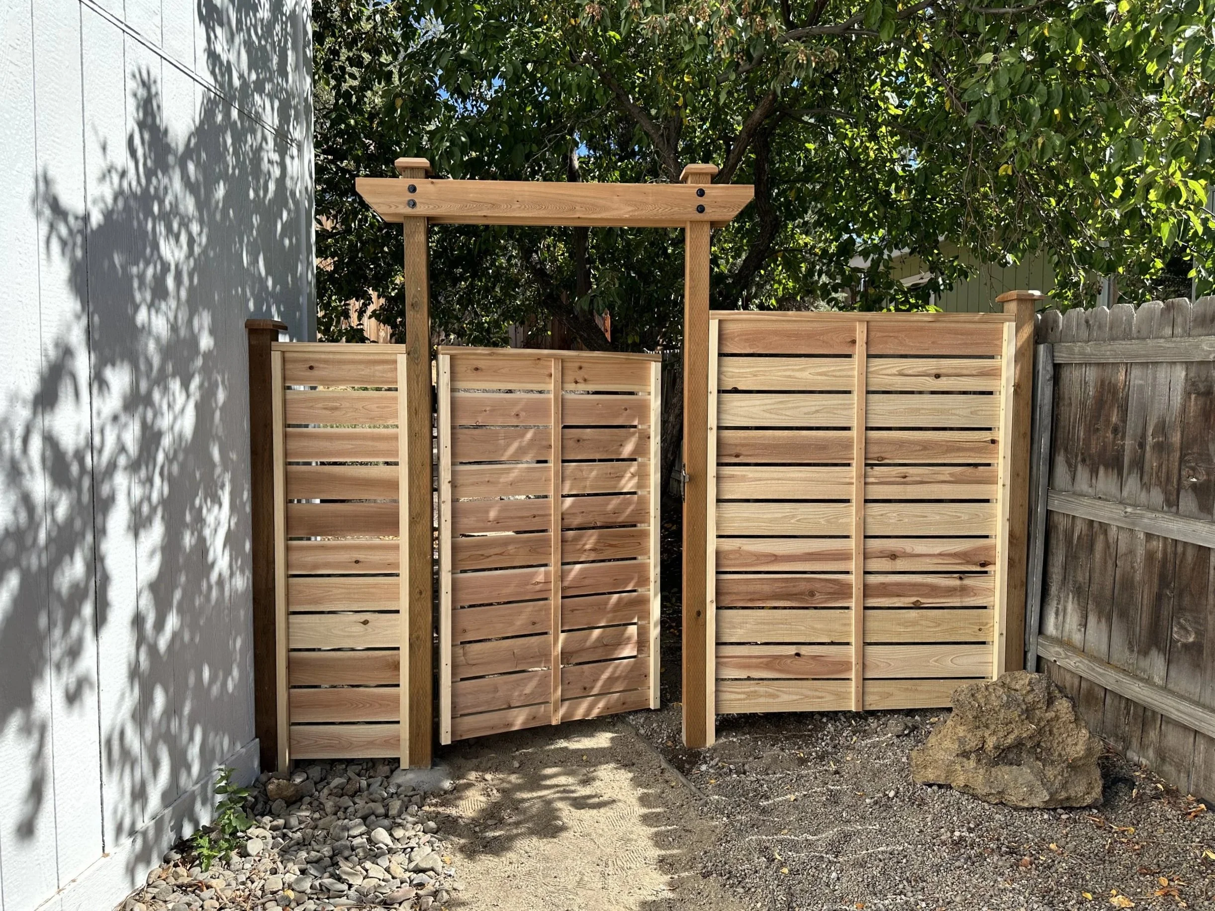 Wooden fence gate with vertical slats and pergola top, surrounded by trees and rocks.