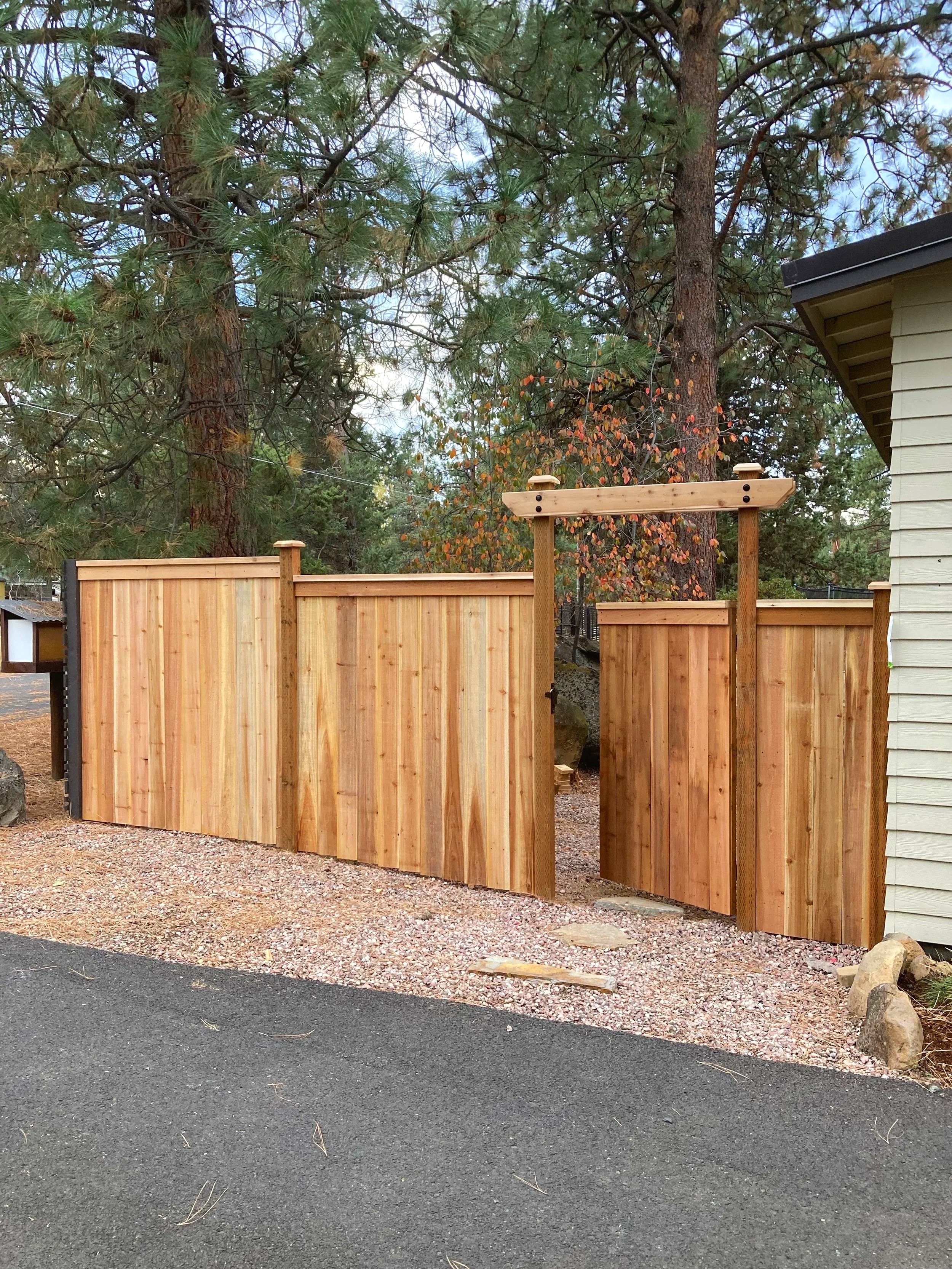 Wooden fence with open gate surrounded by trees and gravel path.