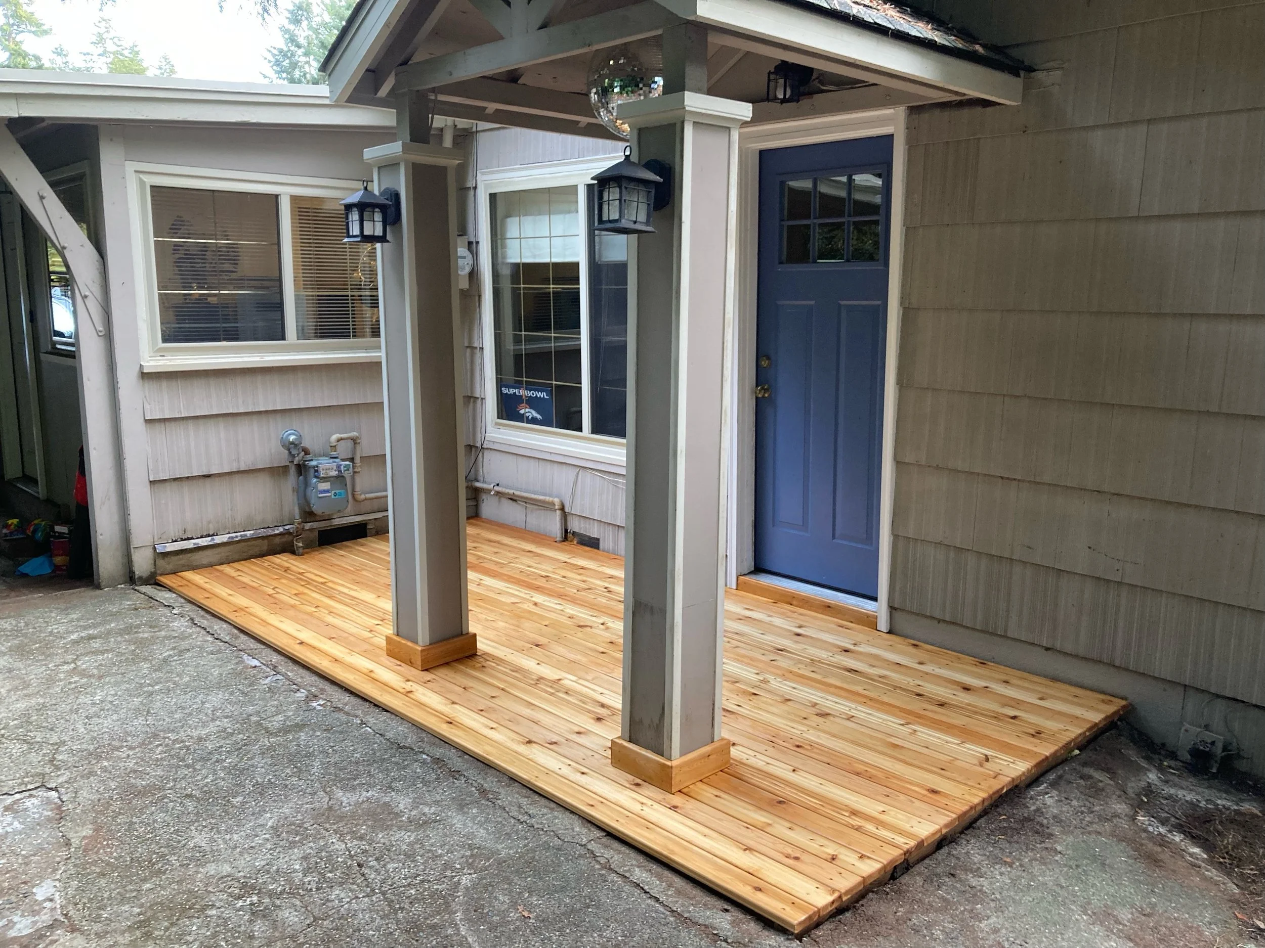 Newly built wooden porch with a blue front door, two black lantern-style lights, and a window with blinds in a beige house, with a concrete surface around.