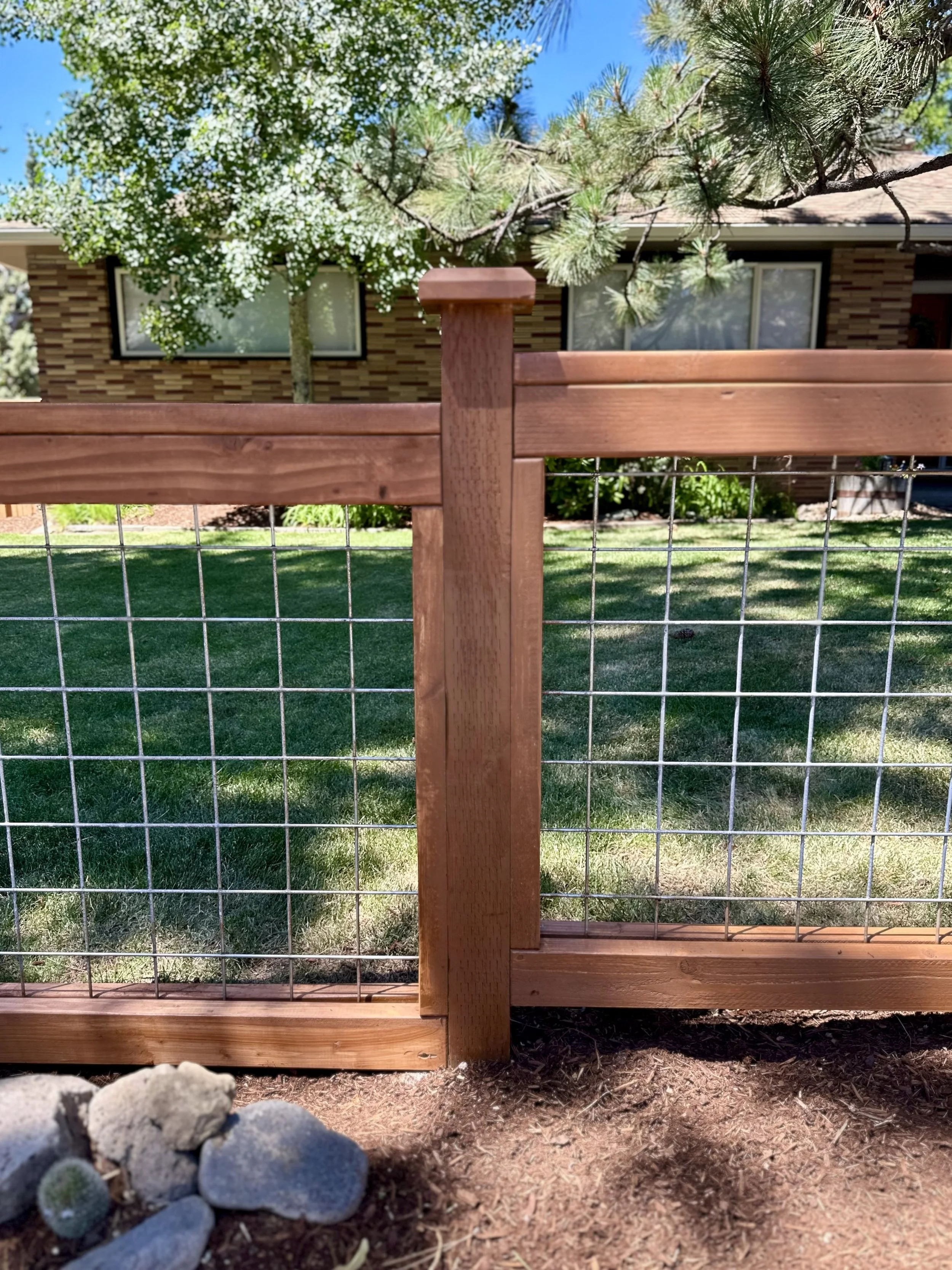 Close-up of a wooden and wire garden fence with a house, trees, and green lawn in the background.