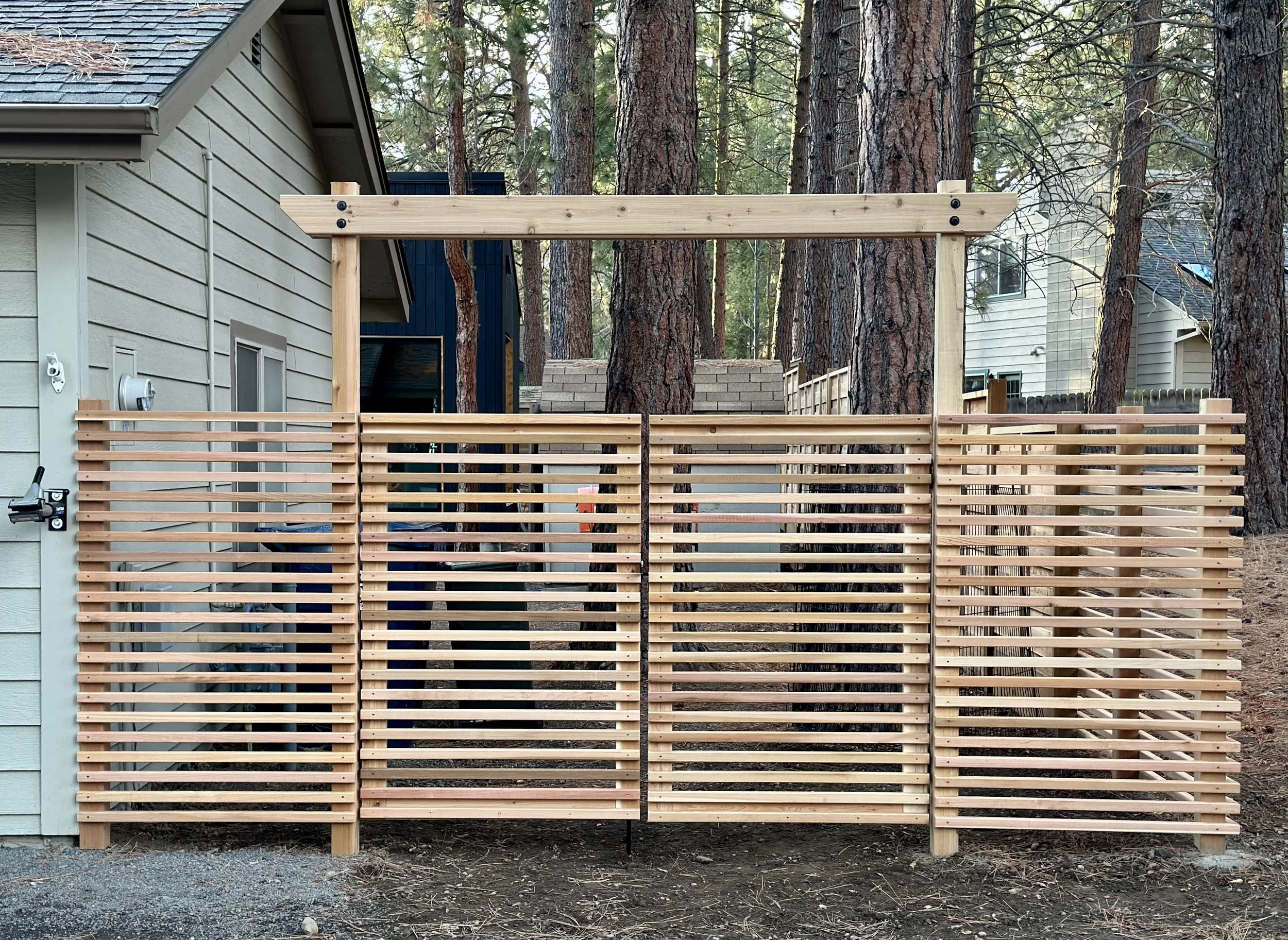 Unfinished wooden fence with horizontal slats in front of a house with light gray siding, set in a wooded area with large pine trees.