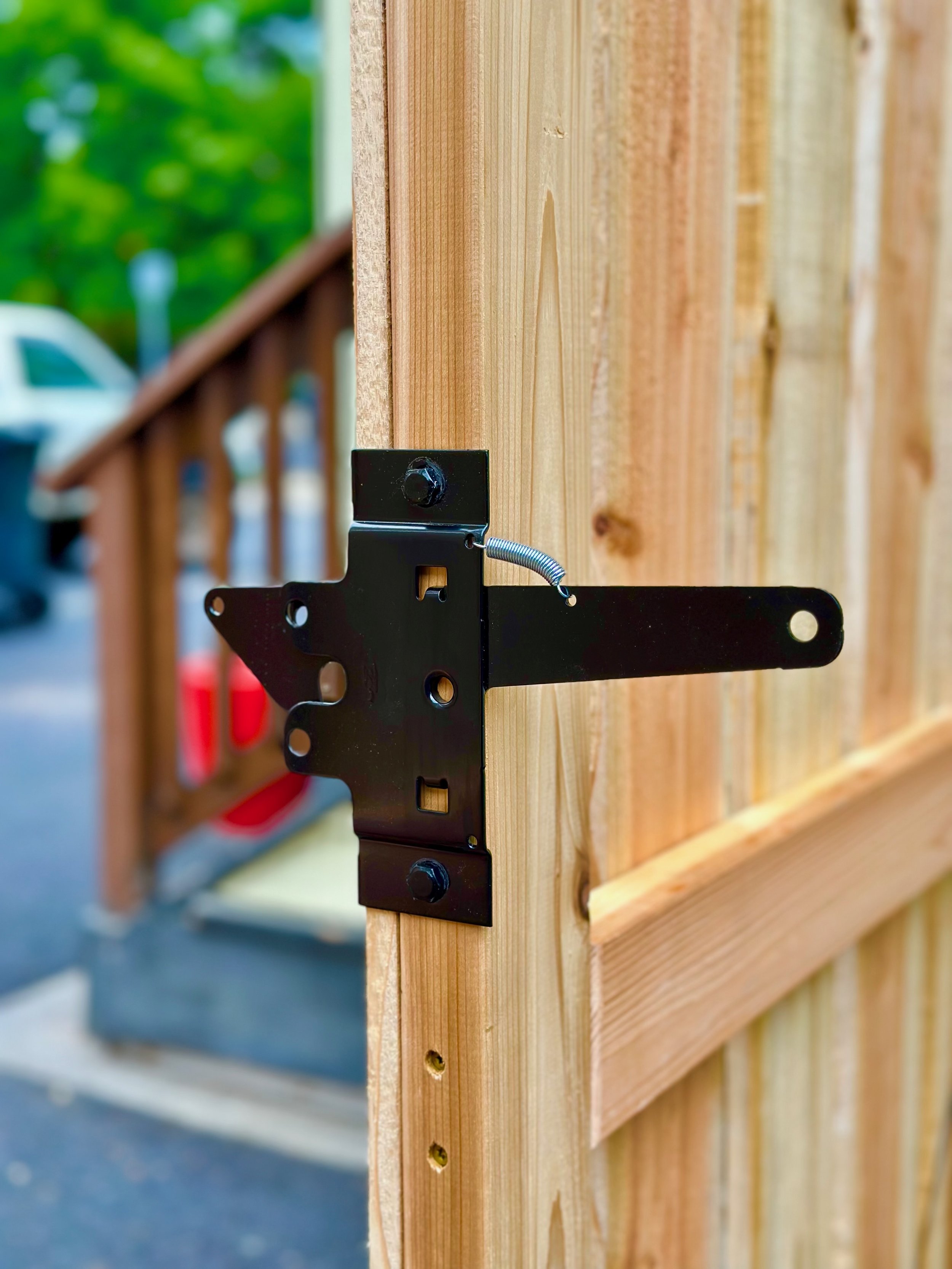 Close-up of a black metal gate latch attached to a wooden fence with blurred background of a truck and trees.