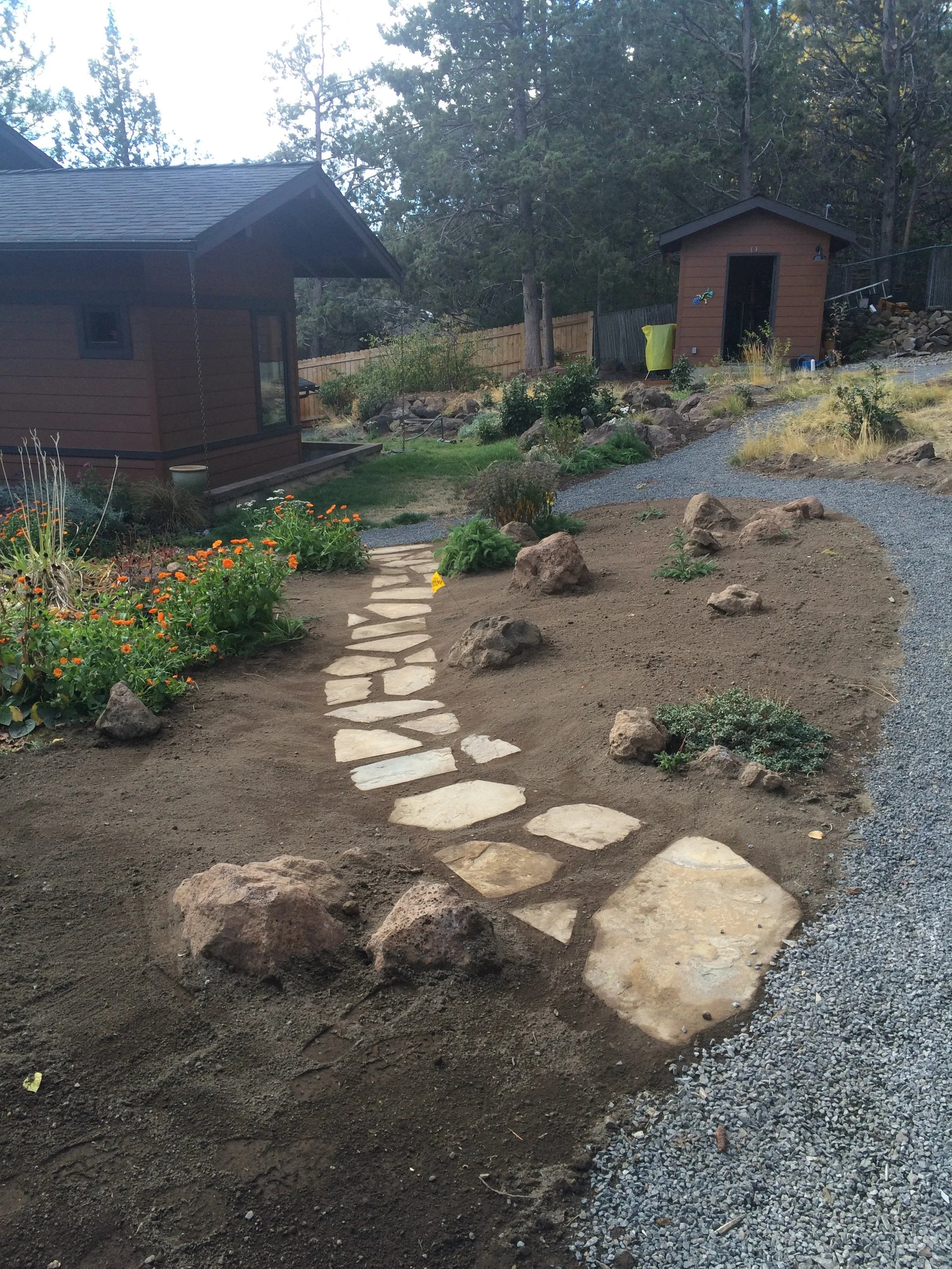 A curved stone pathway in a landscaped yard with flowers, rocks, and two small wooden sheds, surrounded by trees and a wooden fence.