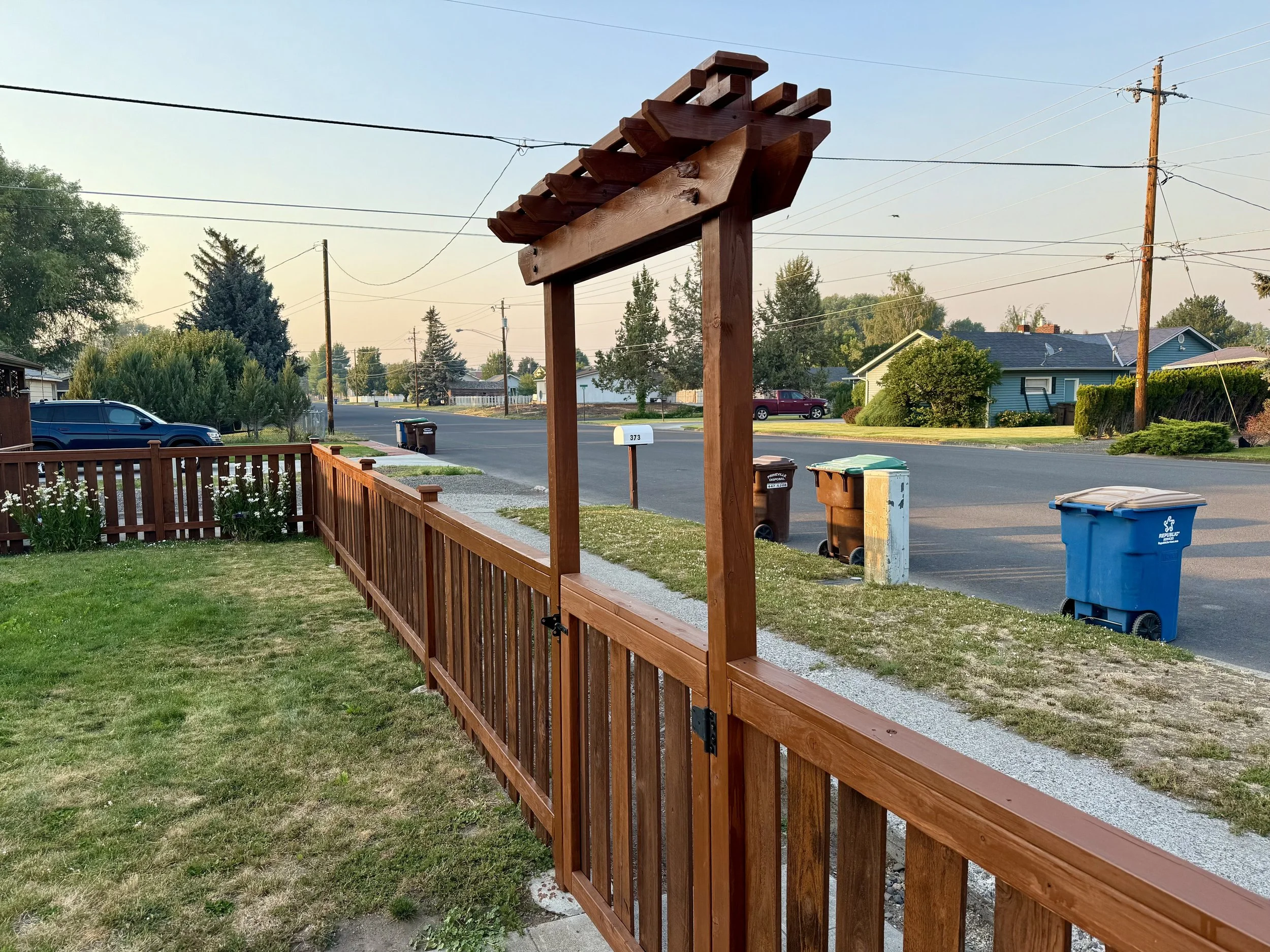 Front yard with wooden fence and gate, street with trash bins, houses, trees, and utility poles in the background during sunset.