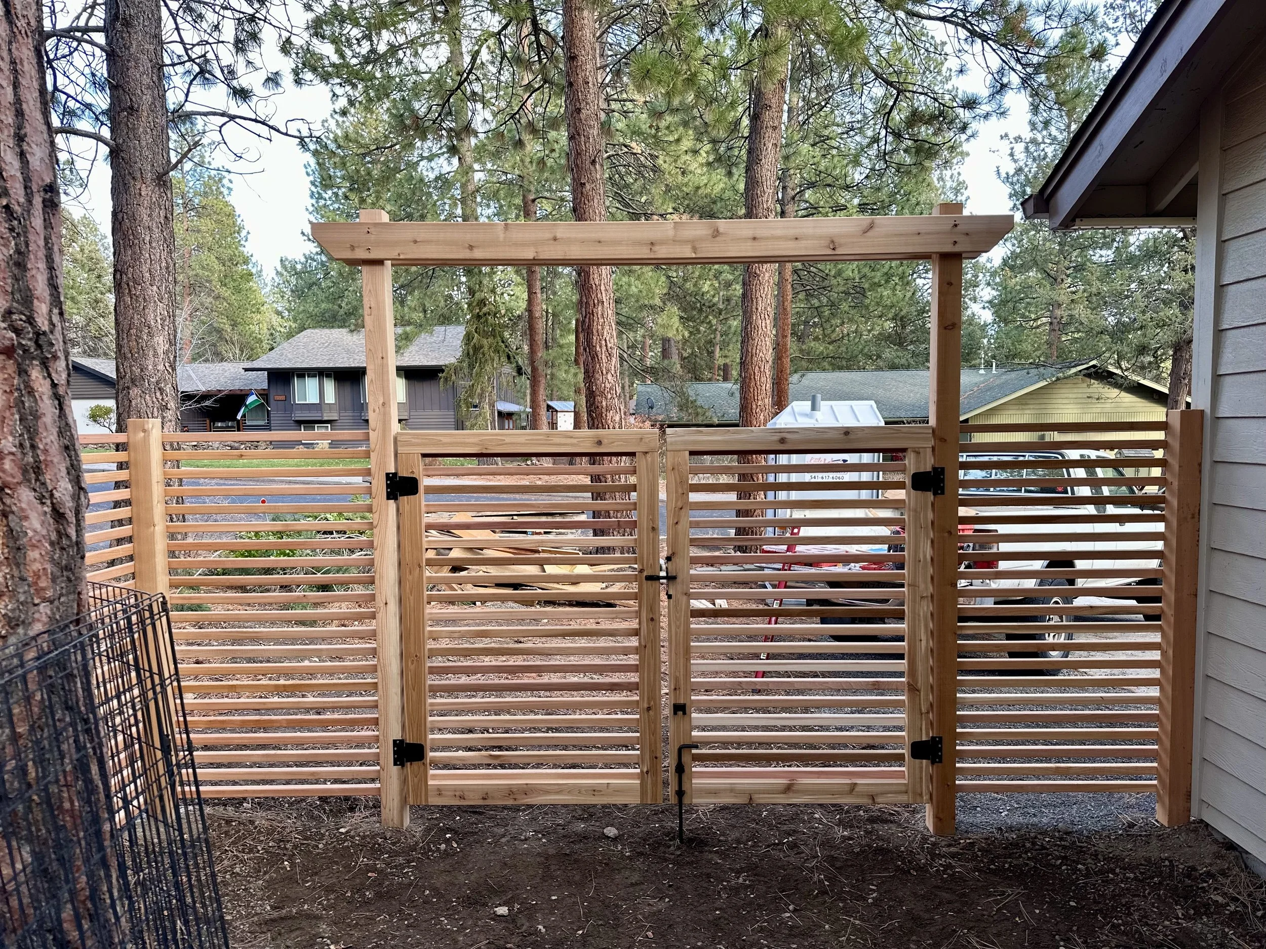 Unfinished wooden gate with horizontal slats and a top frame, situated between a house and a wooded backyard, with trees and neighboring houses in the background.