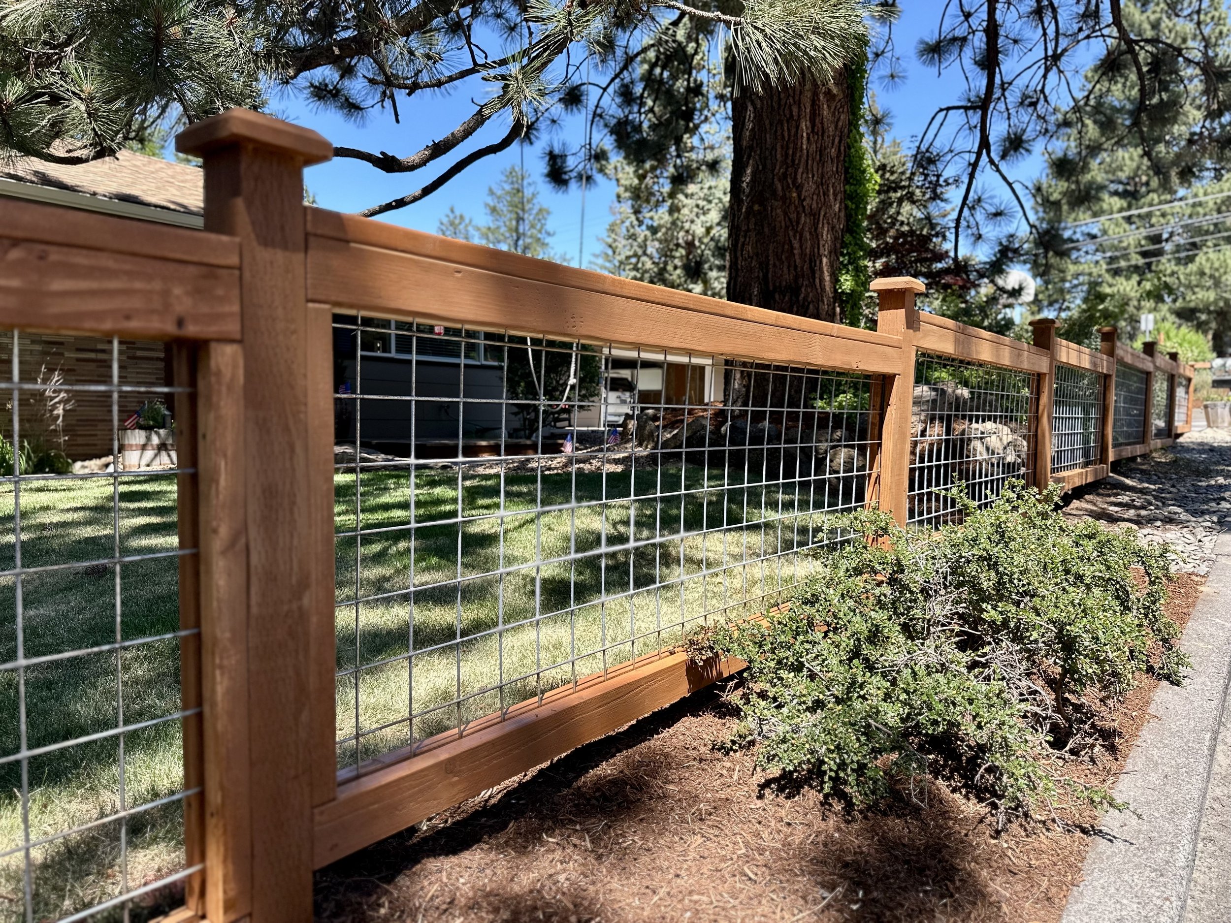 Wooden and wire fence along a sidewalk, with green bushes and trees in the background under a clear blue sky.