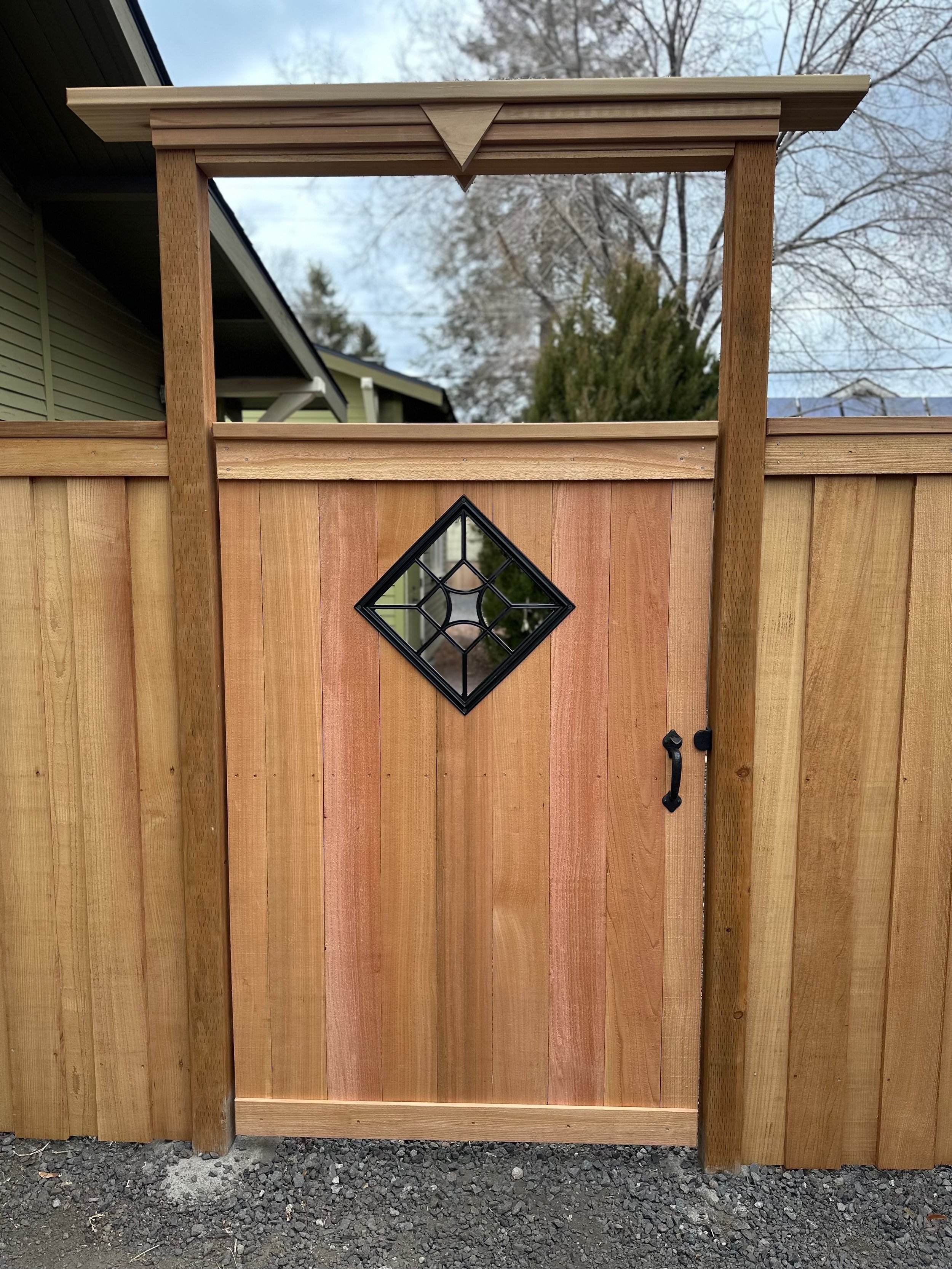 Close-up of a wooden gate with a diamond-shaped window and a small roof extension on top, attached to a wooden fence.