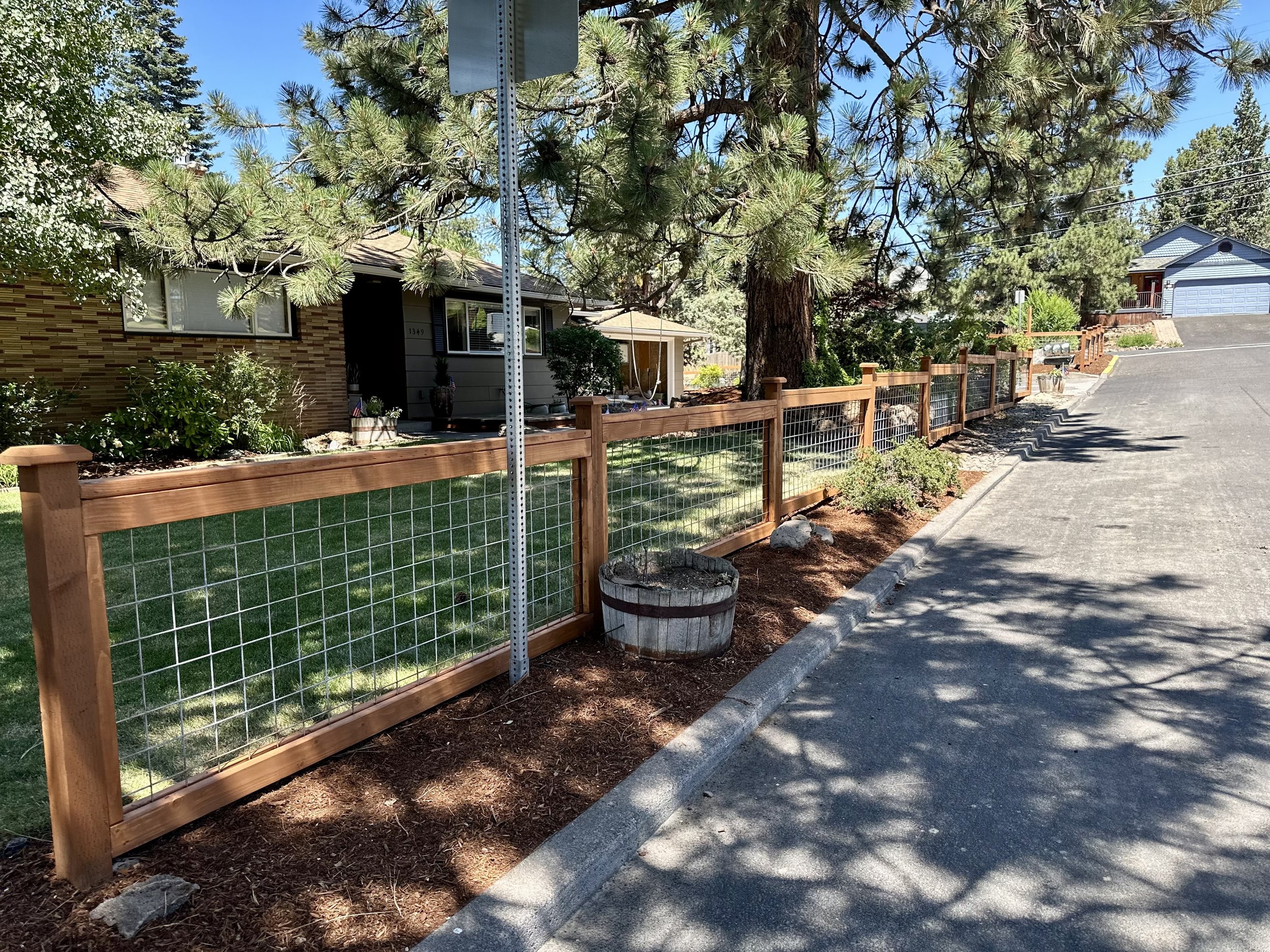 A residential front yard with a wooden fence, green grass, shrubs, large tree, potted plants, and neighboring houses on a clear day.