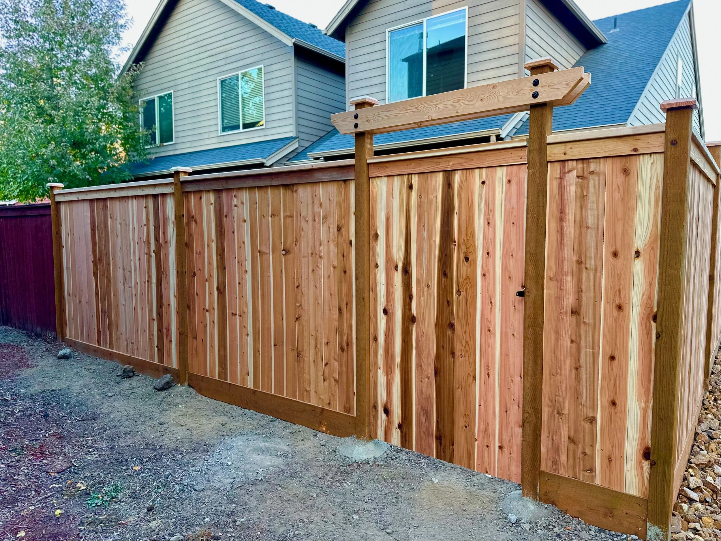 A newly built wooden fence with vertical planks and horizontal support beams in front of a modern multi-story house with beige siding and blue roof.