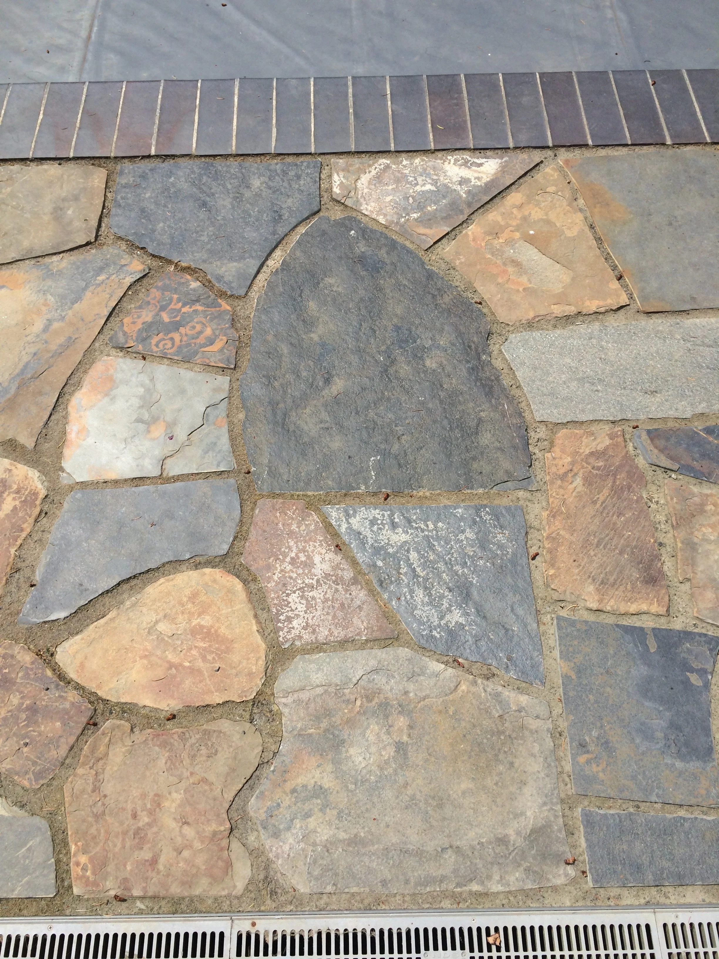 Close-up of a stone patio with various irregularly shaped stones and a metal drainage grate at the bottom.