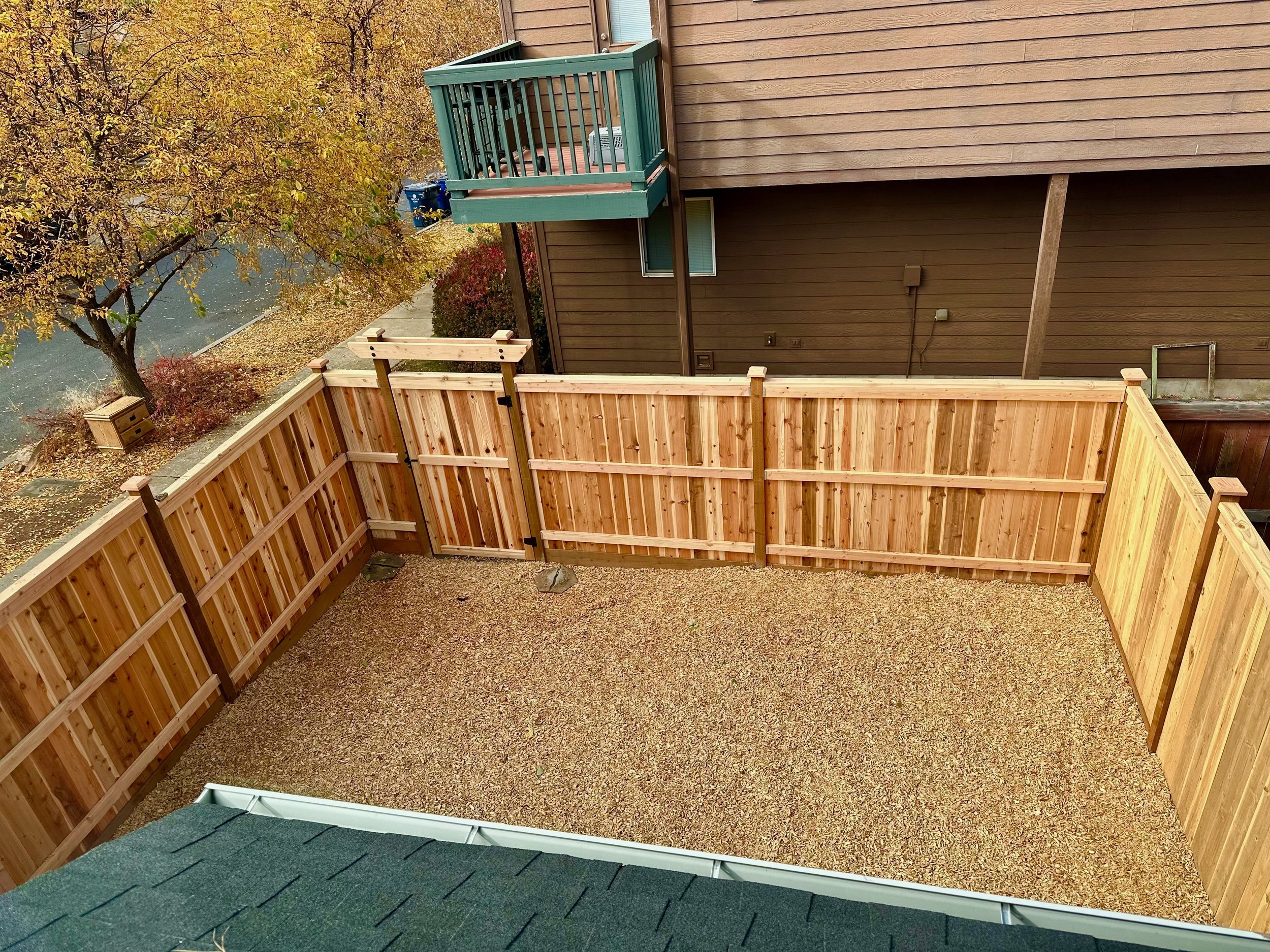 View of a fenced backyard with a gravel ground, wooden fence, neighboring house with a teal balcony, and trees with autumn-colored leaves.