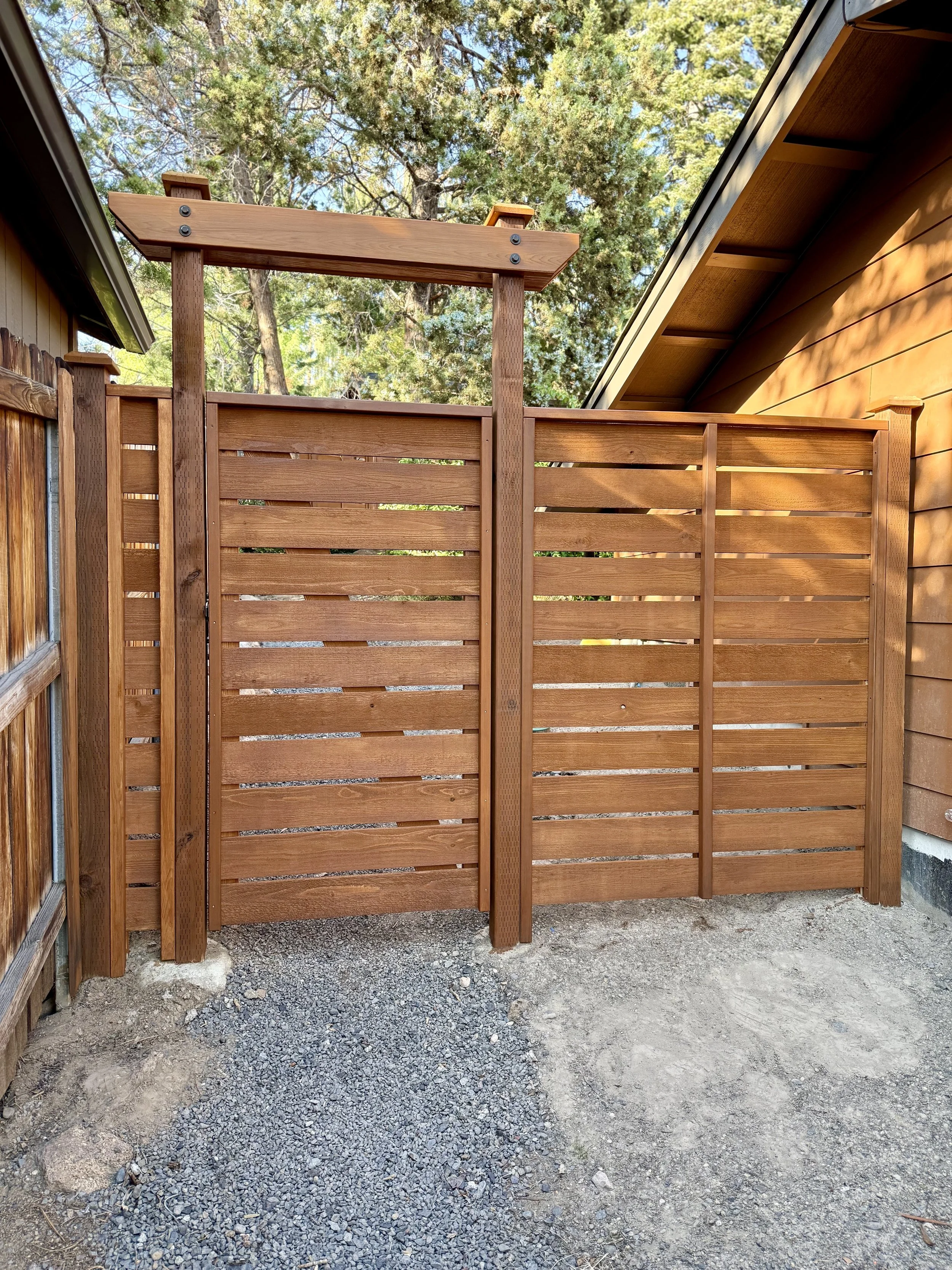 A wooden privacy fence with horizontal slats and vertical posts, set against a background of trees and a building with brown siding.
